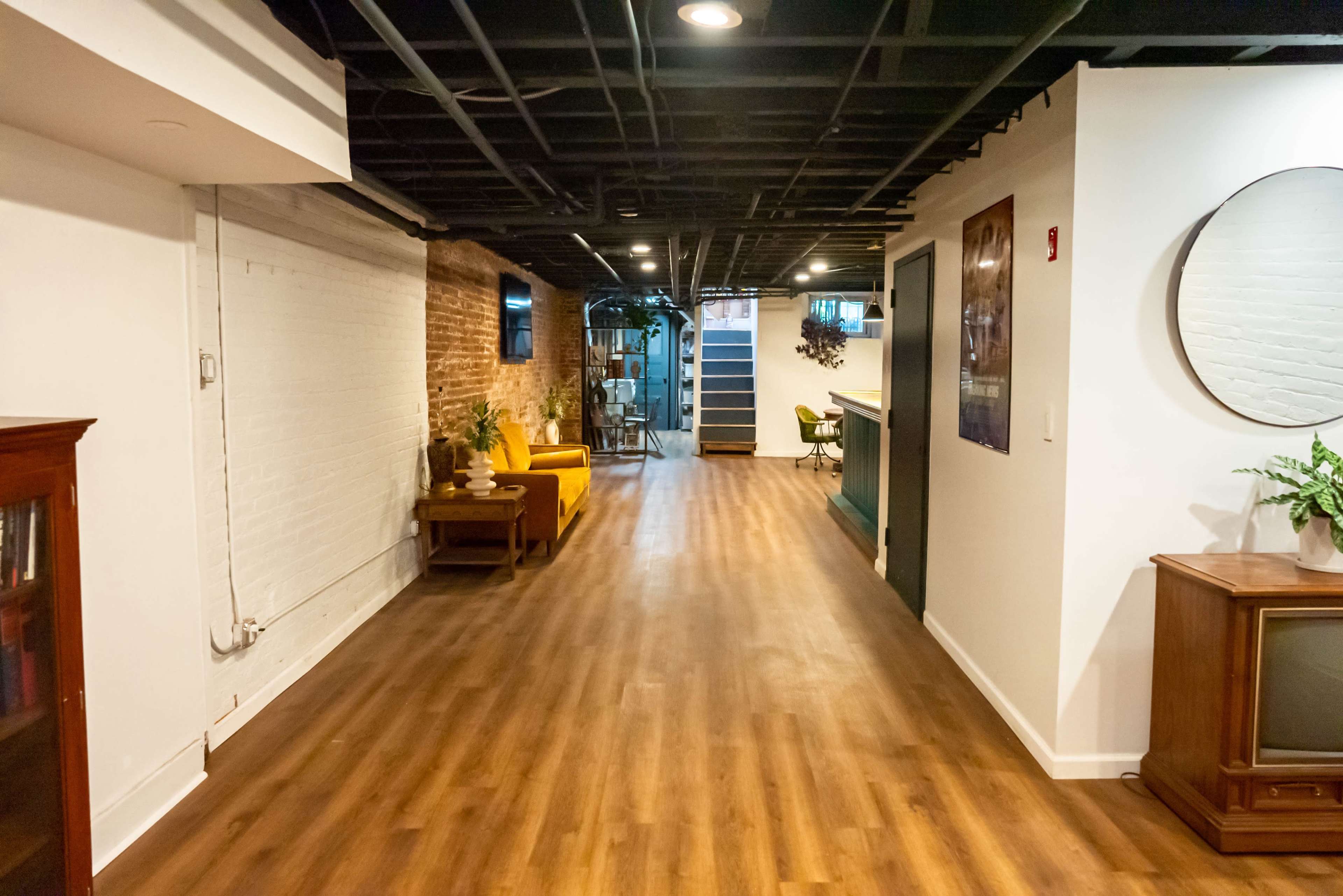 A long hallway with wooden flooring, leading to a staircase and featuring a sitting area with yellow chairs and a vintage TV.