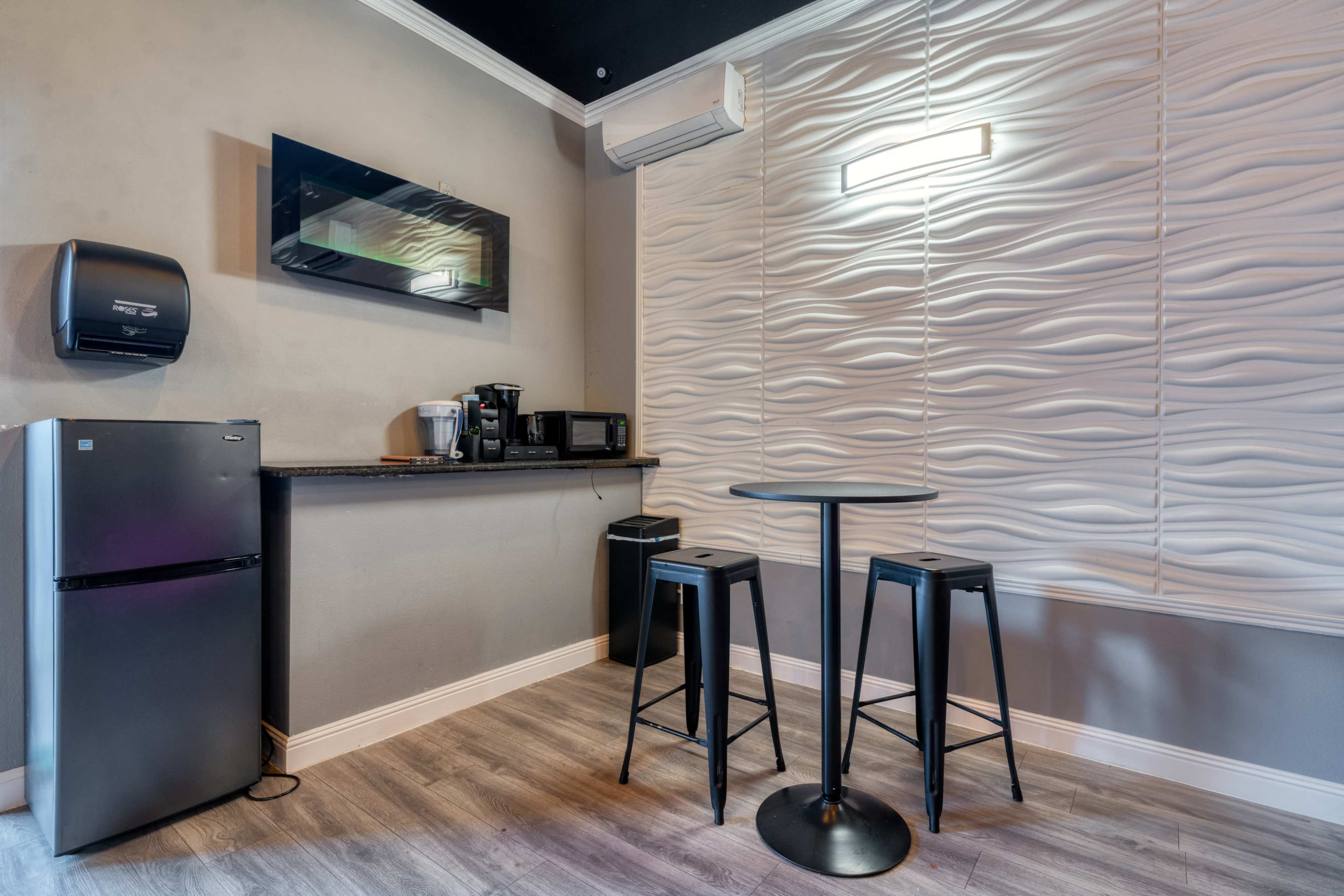 A compact kitchen area featuring a small round table with two stools, a refrigerator, a coffee maker, and a microwave against a textured wall.