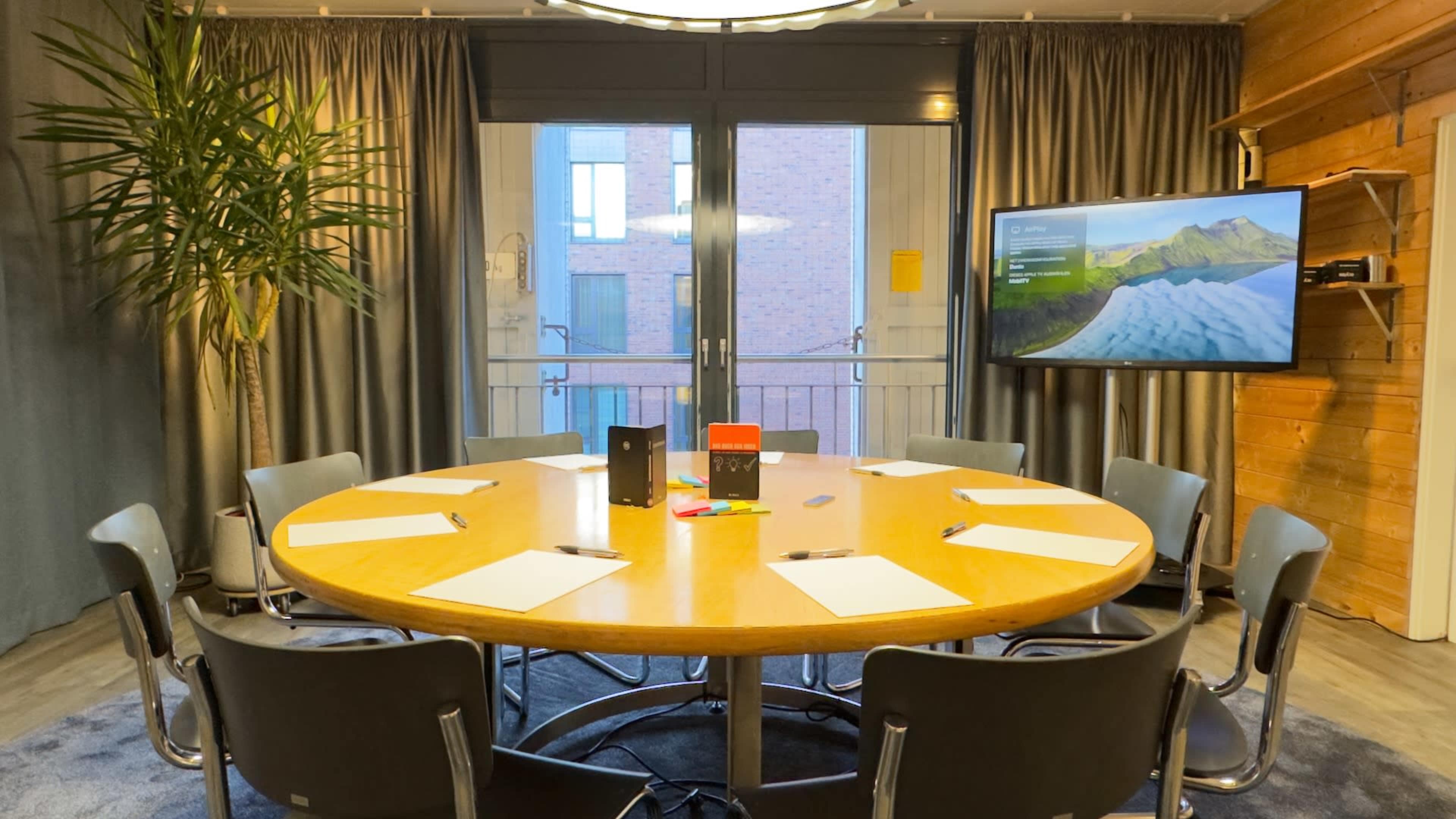 A round wooden table surrounded by eight chairs is set up in a conference room with large windows and a screen displaying a landscape image.