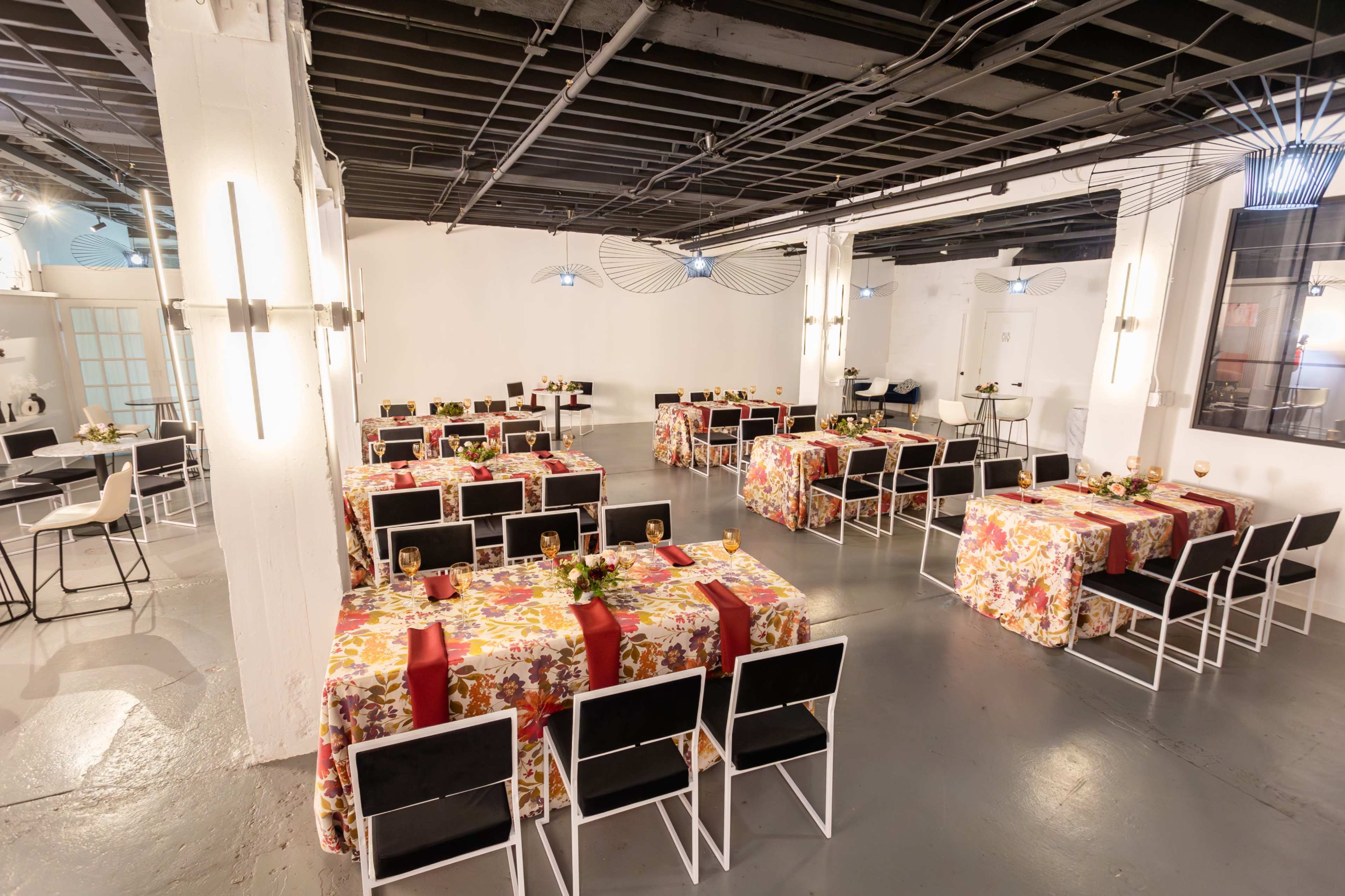 A spacious event venue features several tables covered with floral tablecloths, arranged in a grid pattern with black chairs.