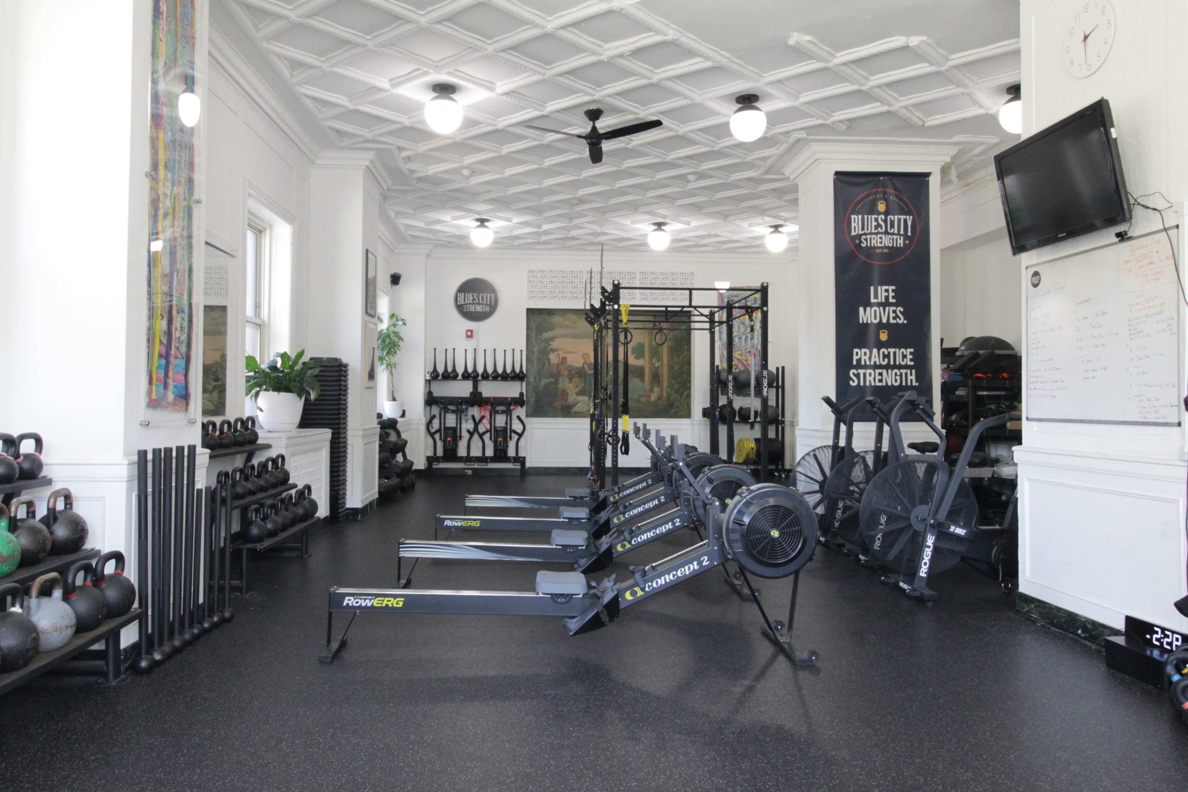 The image shows a fitness center equipped with rowing machines, weights, and a pull-up rig under a patterned ceiling.