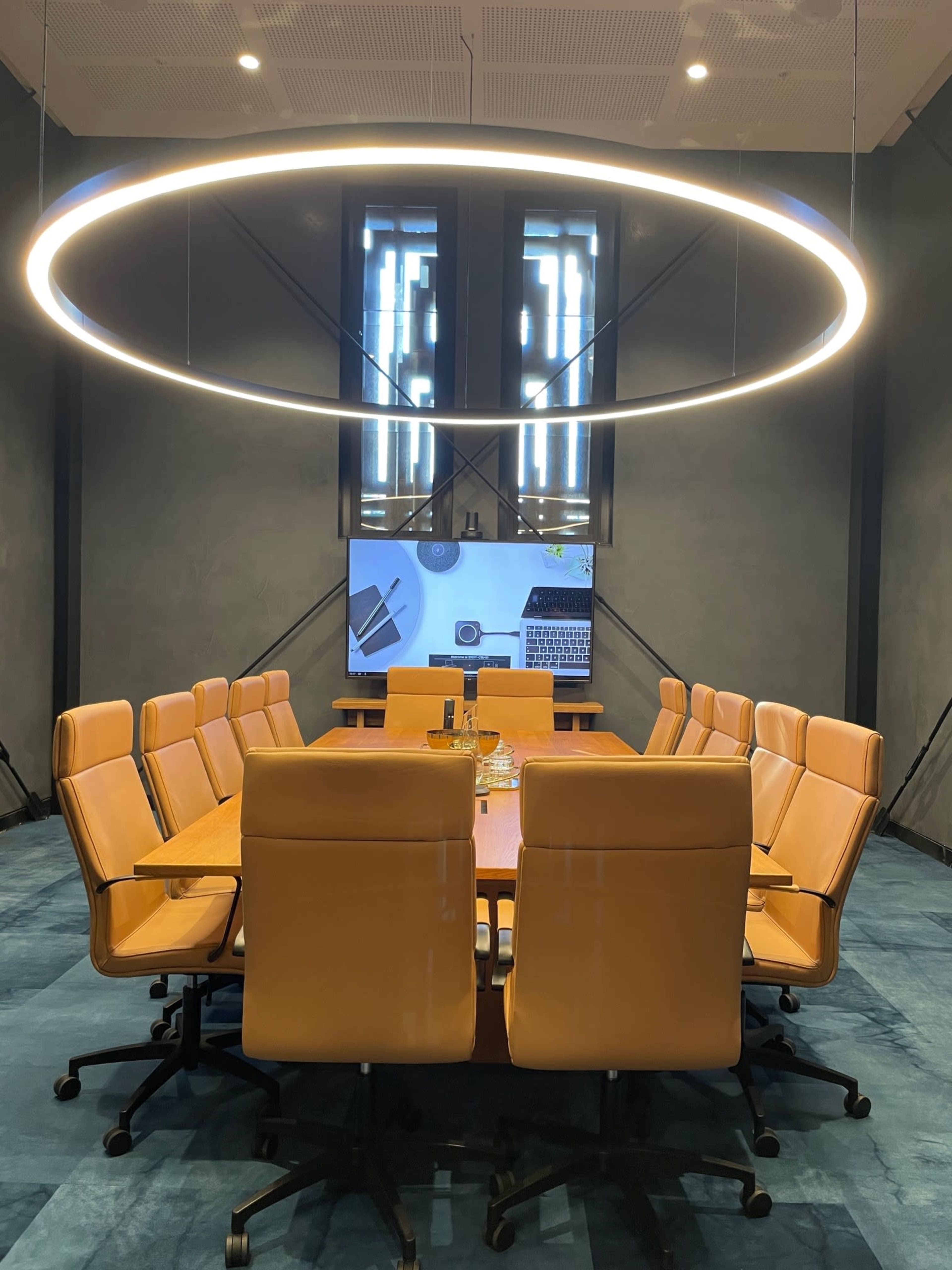 A modern conference room featuring a large wooden table surrounded by orange chairs and a circular light fixture overhead, with a screen displaying a laptop and office-related items.