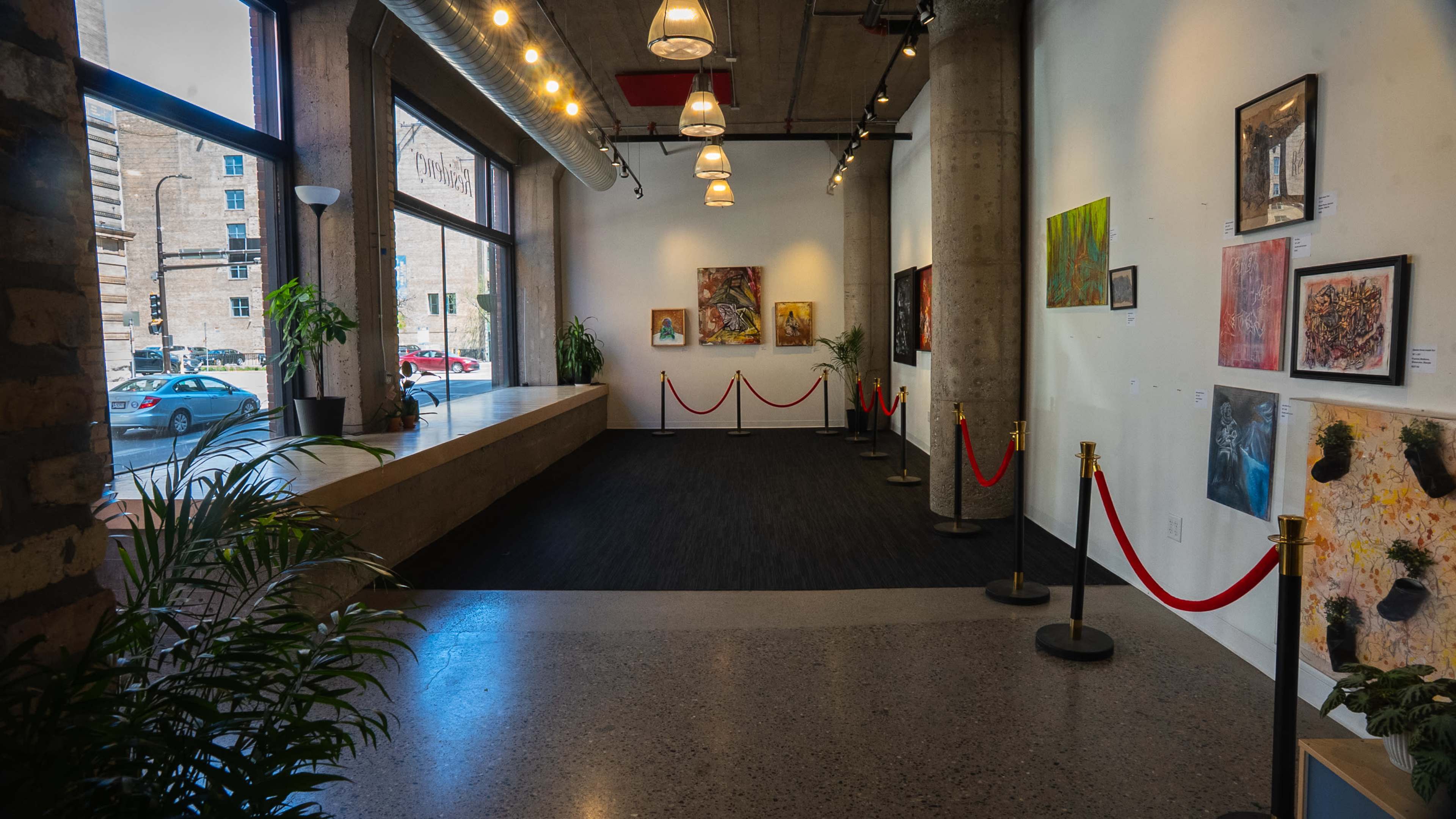 The image shows an art gallery space with various artworks displayed on walls, red ropes guiding the visitor path, and large windows allowing natural light to fill the room.