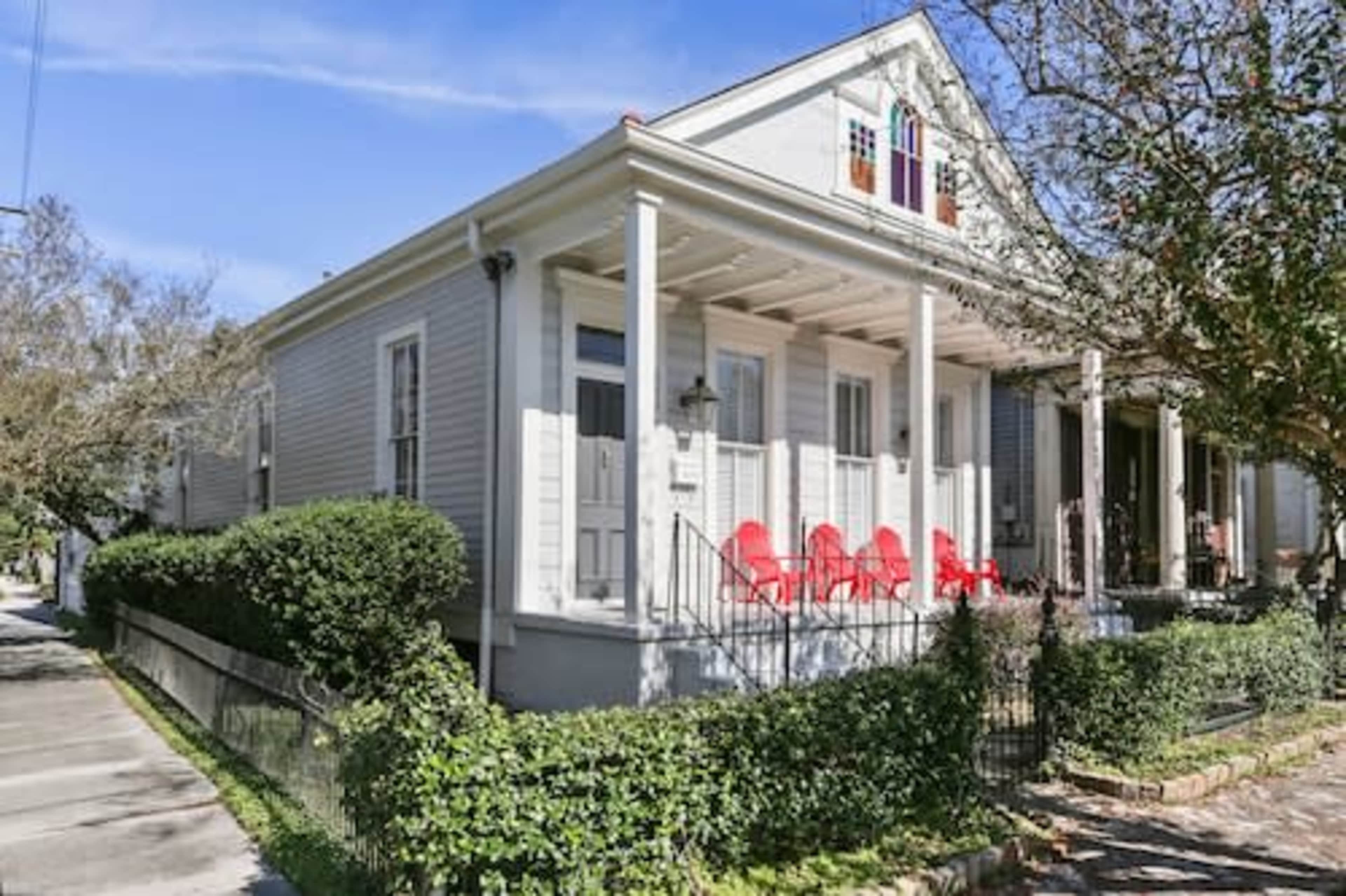 A light-colored house with a covered porch and red rocking chairs sits at the edge of a well-maintained garden.