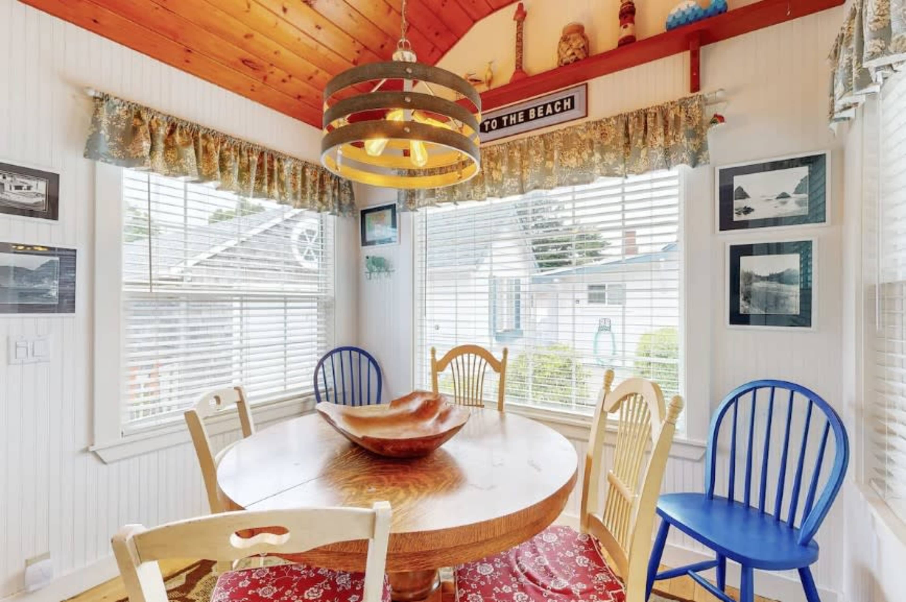 A round wooden table is set in a brightly lit dining area with blue and white chairs, surrounded by large windows adorned with patterned curtains and framed photographs on the walls.