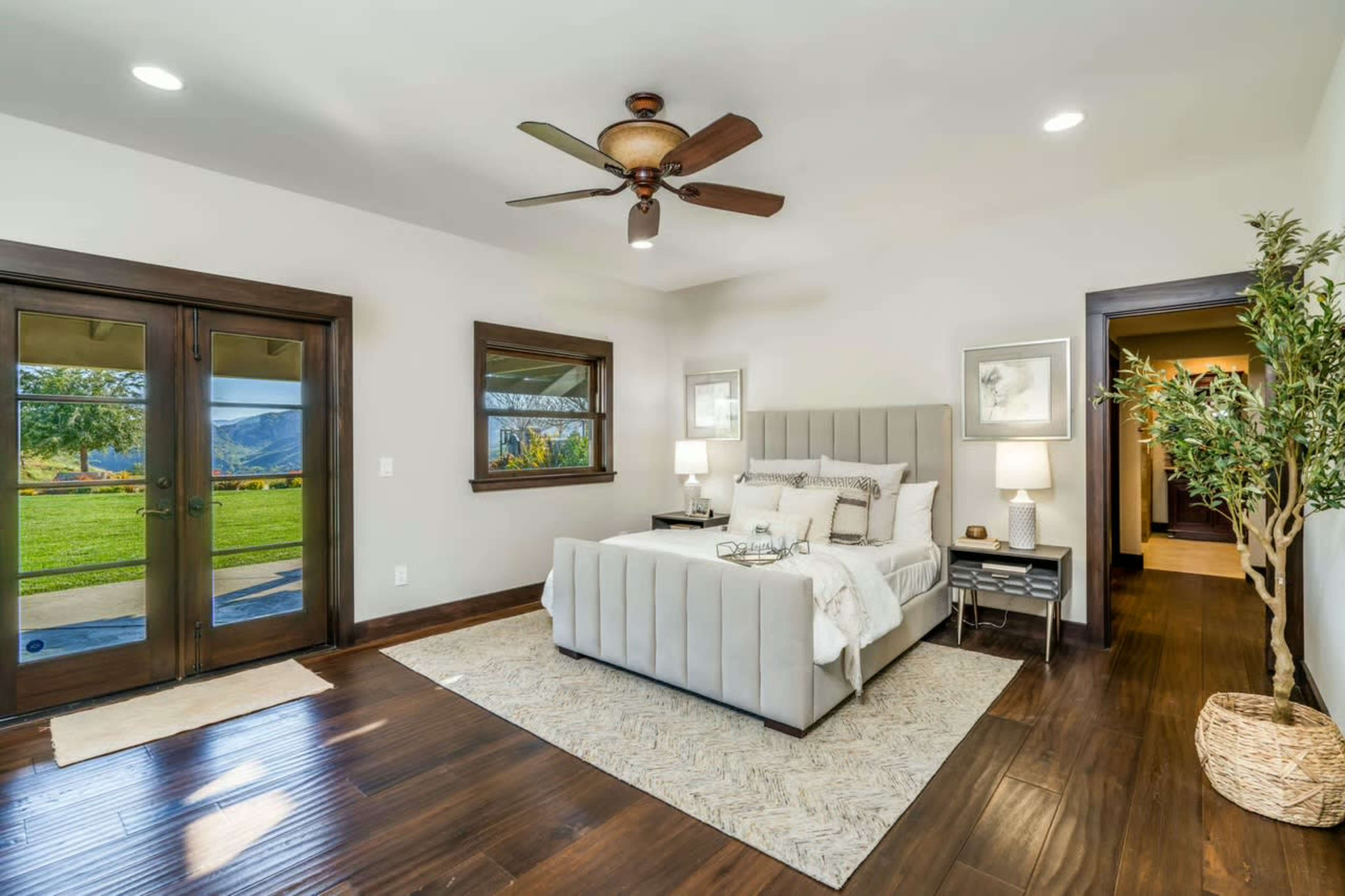The image shows a spacious bedroom with a king-sized bed, wooden flooring, and large windows leading to a green outdoor view.