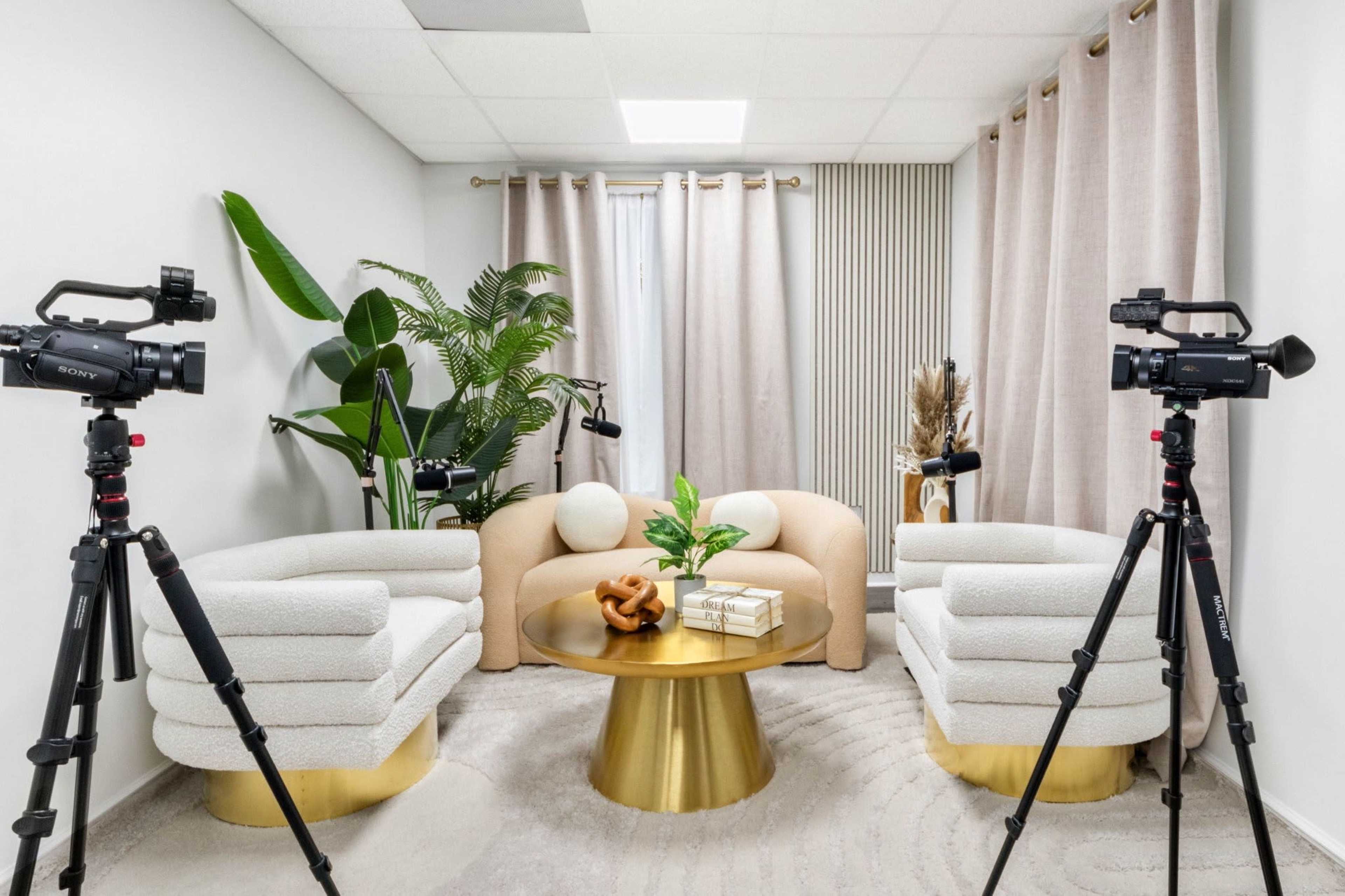The image shows a modern interview setup with two cameras positioned on tripods facing a cozy seating area featuring beige sofas, a gold circular coffee table, and decorative plants.