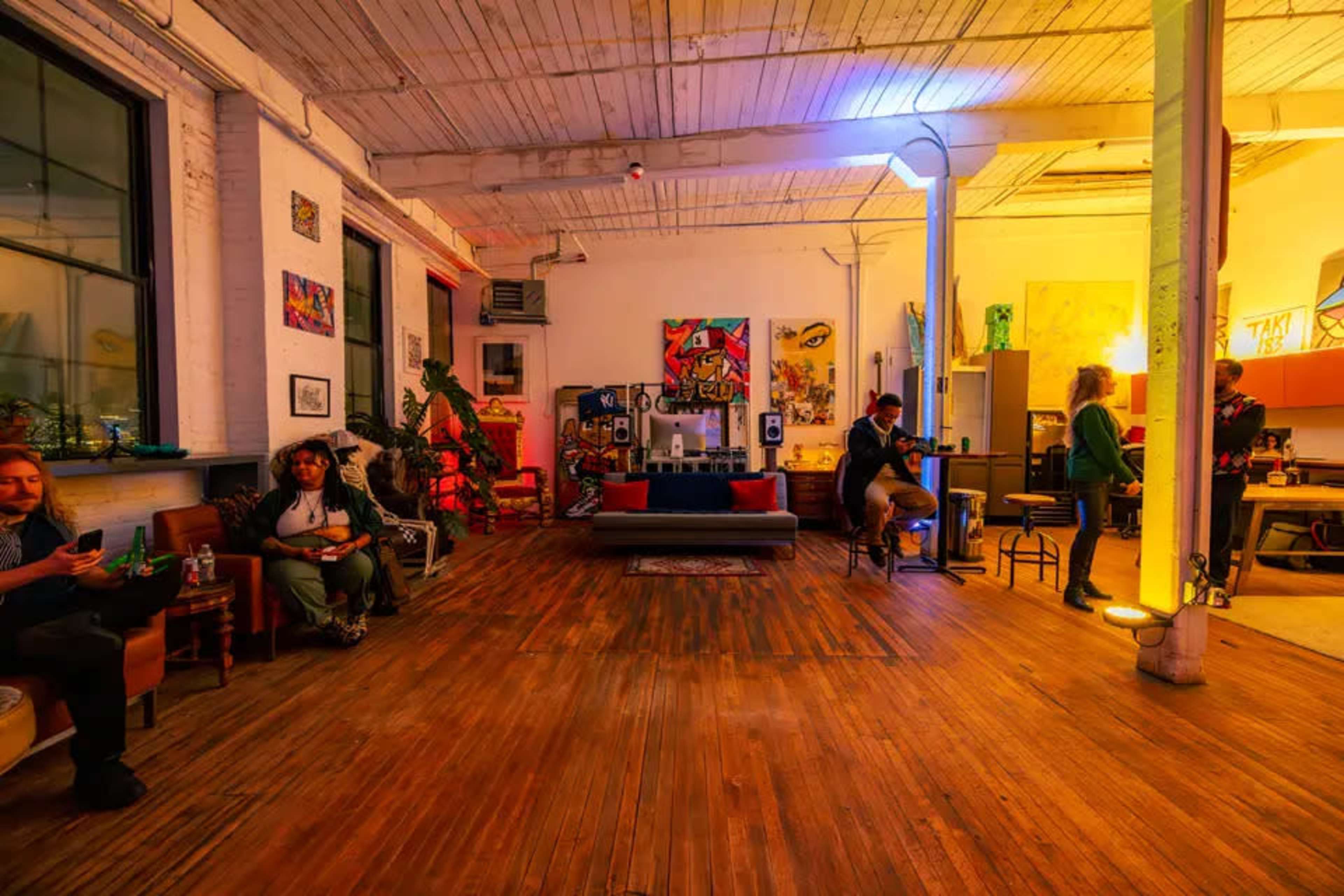 The image shows a spacious, well-lit room with wooden floors, featuring various artworks on the walls and several people engaged in different activities.