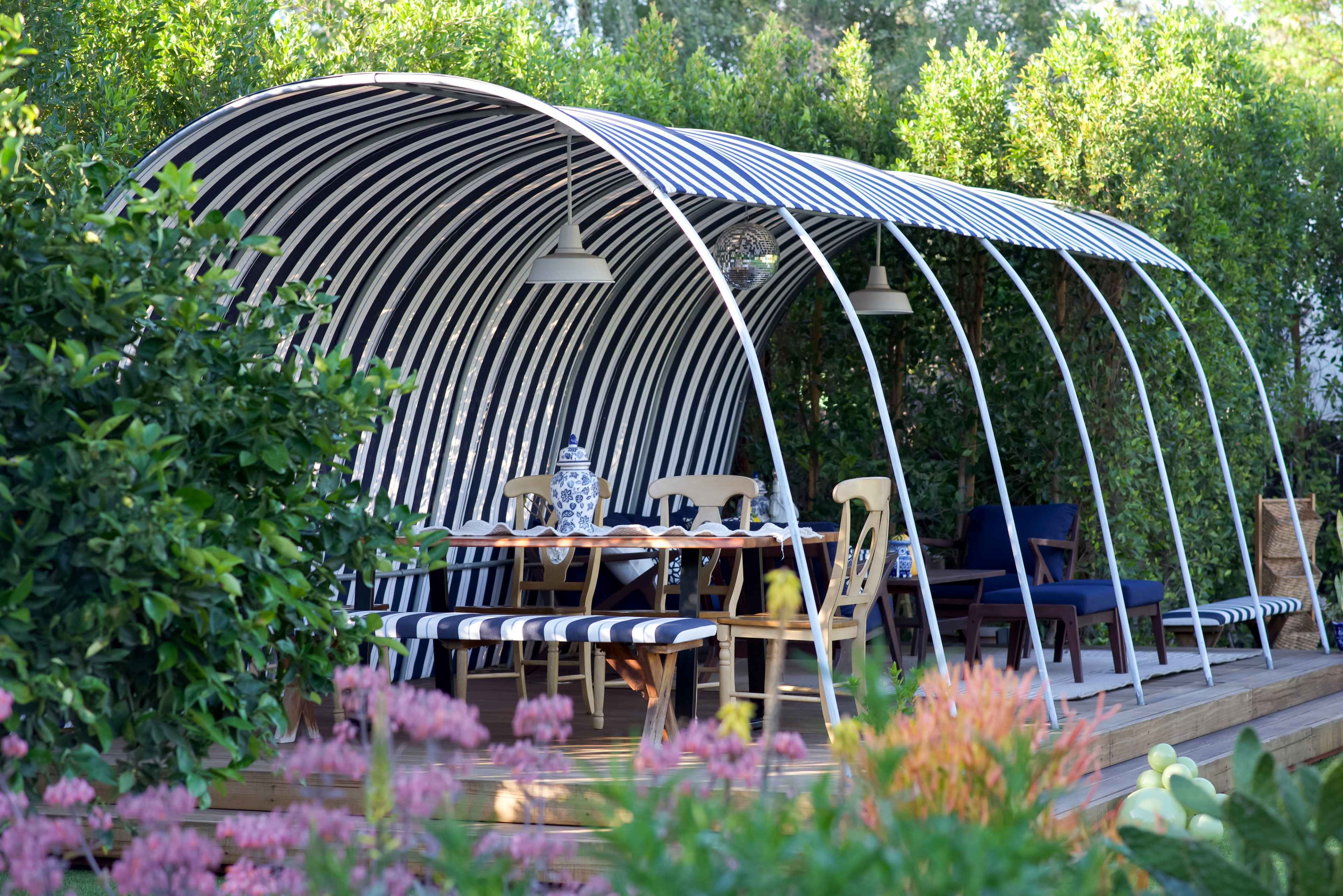 A modern outdoor dining area features a curved striped canopy, wooden deck, and a table set with chairs, surrounded by lush greenery.