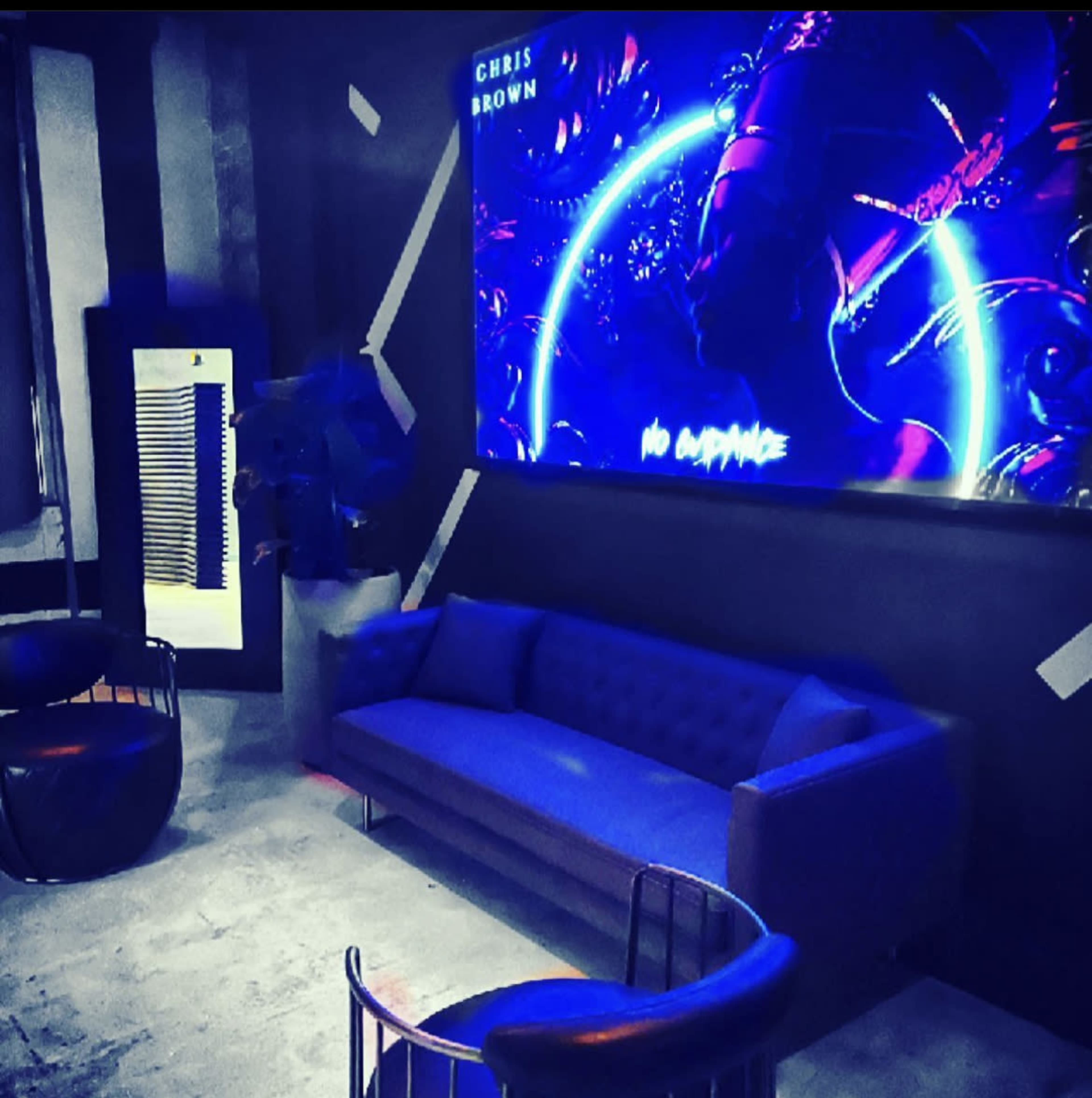 A modern lounge area with a blue couch and chairs, illuminated by a large screen displaying vibrant graphics and the text "NO GUIDANCE."