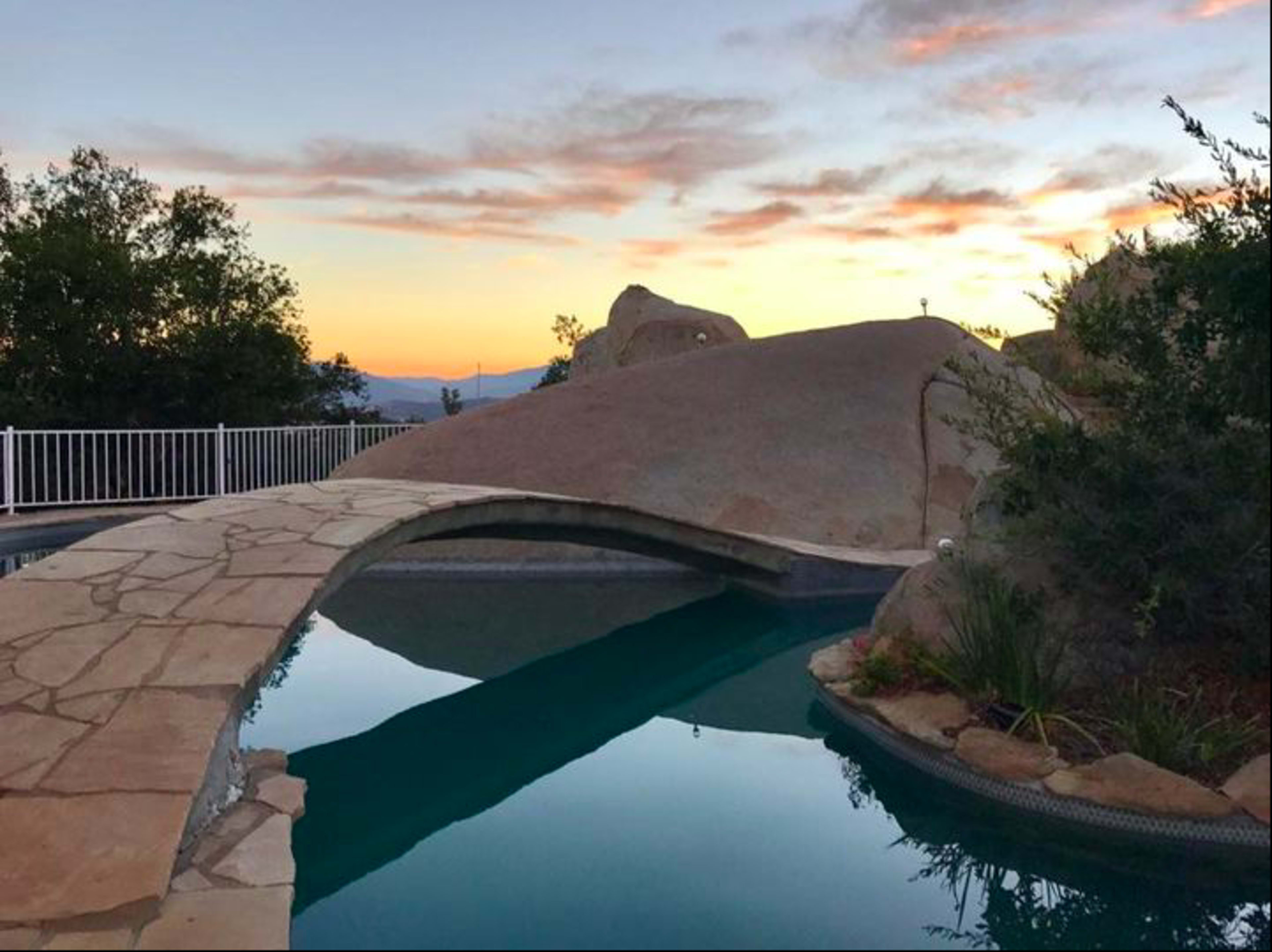 A stone pathway arches over a swimming pool, leading to a rocky landscape under a colorful sunset sky.