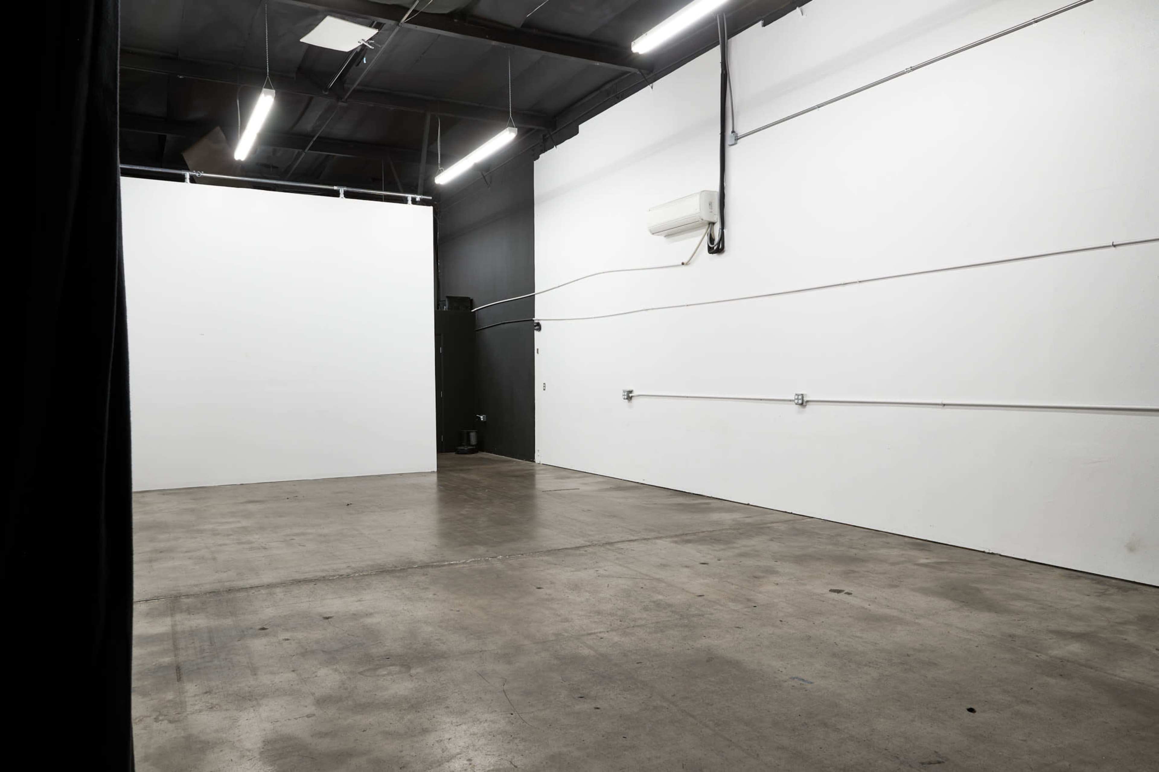 An empty, industrial-style room with concrete flooring and white walls, featuring overhead lighting and exposed electrical elements.