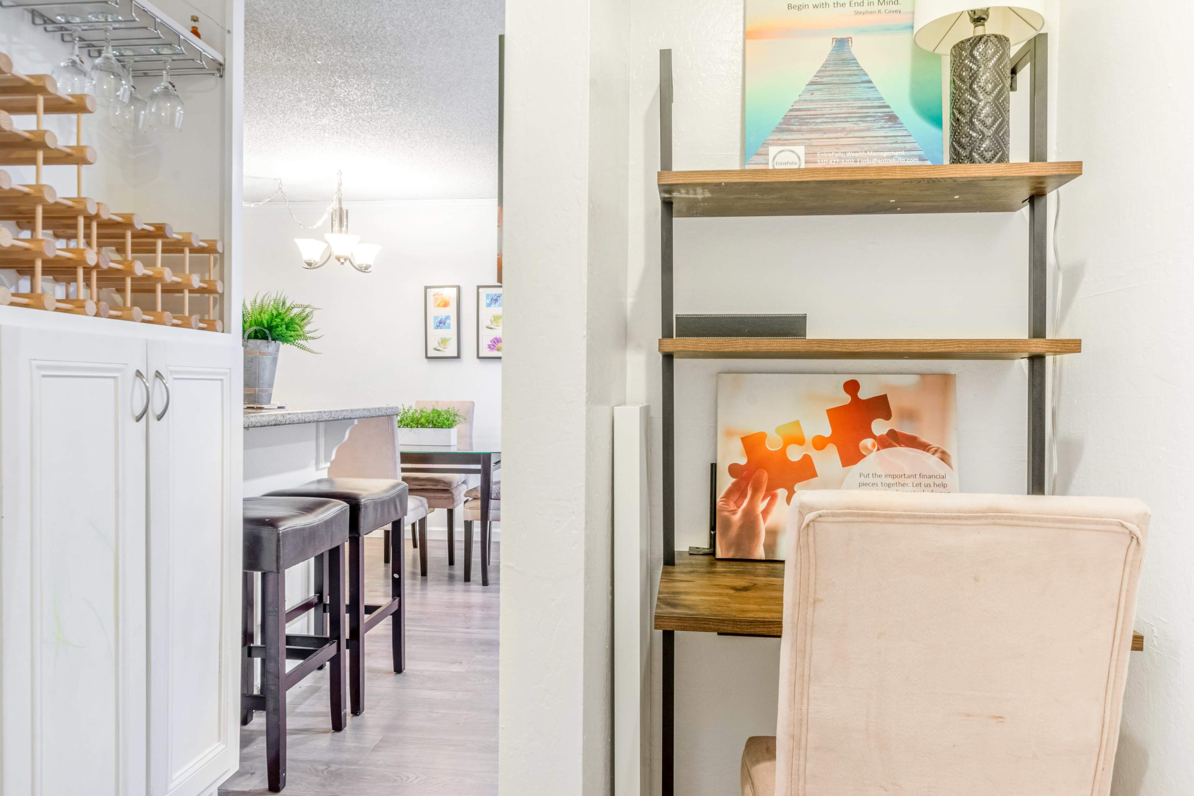 The image shows a modern living space featuring a bookshelf with decorative items and a light-colored chair, with a view of a kitchen area in the background.
