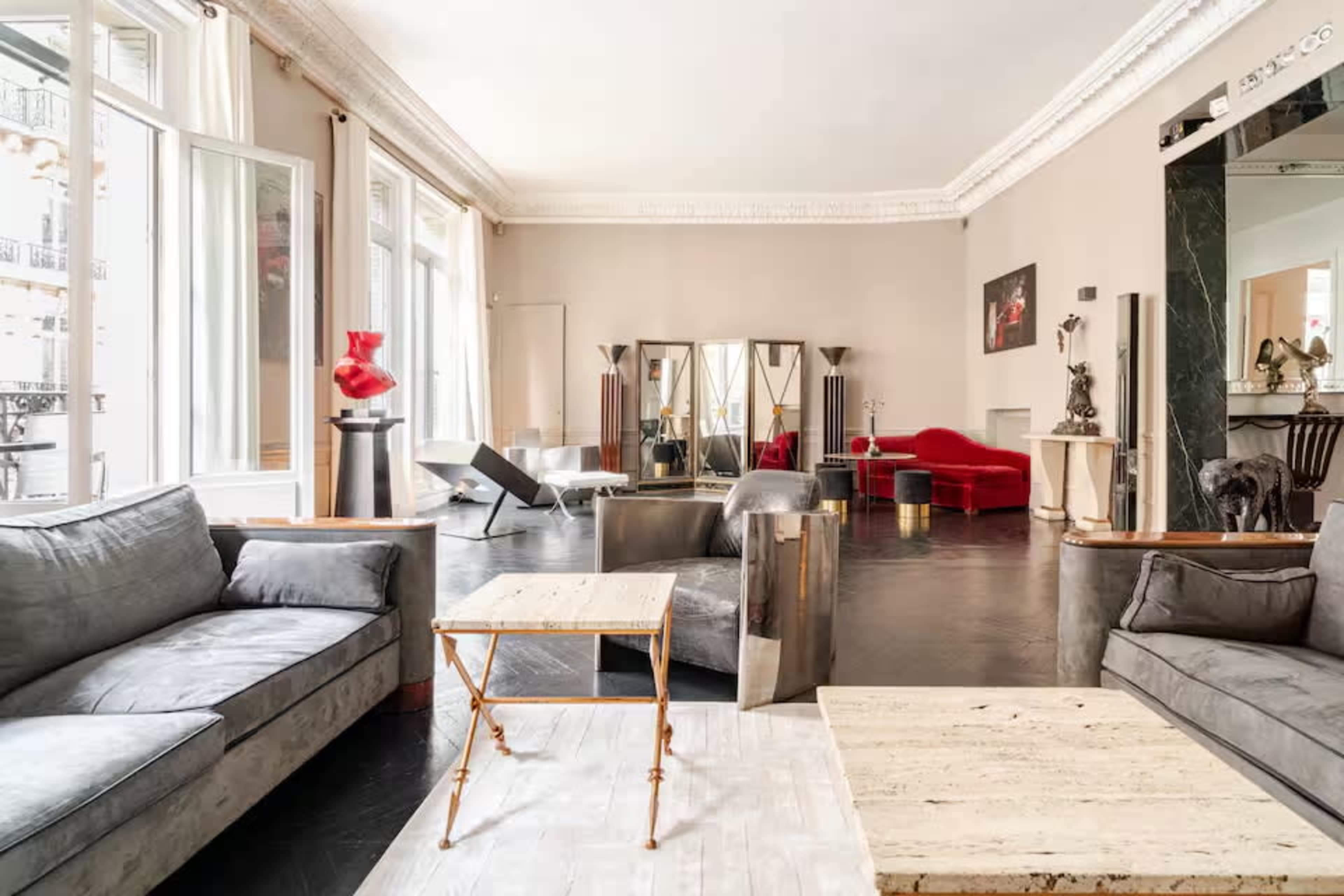 The image shows a spacious living room with a mix of modern furniture, including a red sofa, a minimalist coffee table, and large windows allowing natural light.