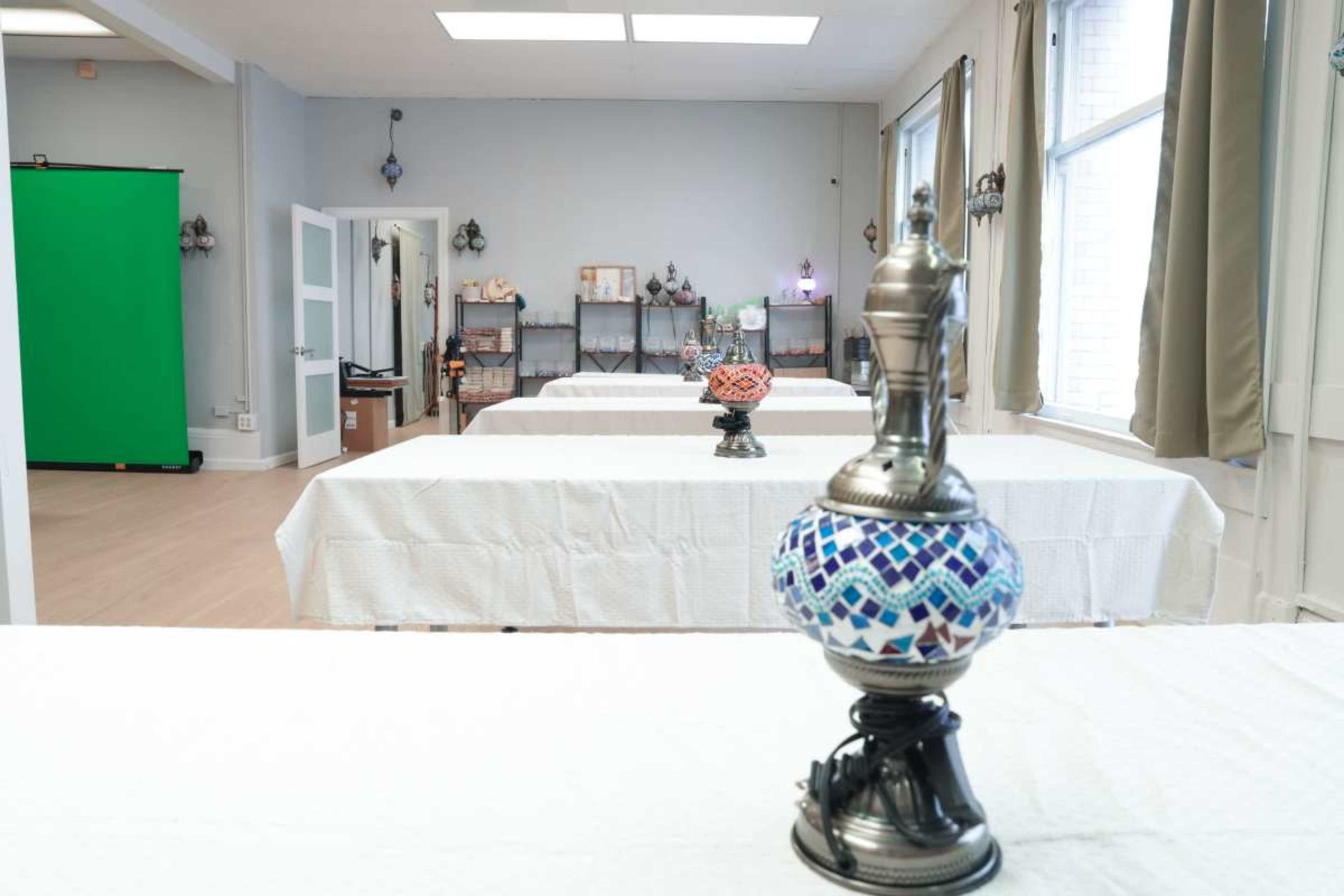 A decorative lamp is positioned in the foreground, illuminating a spacious room with multiple tables covered by white tablecloths and a green screen at the far end.