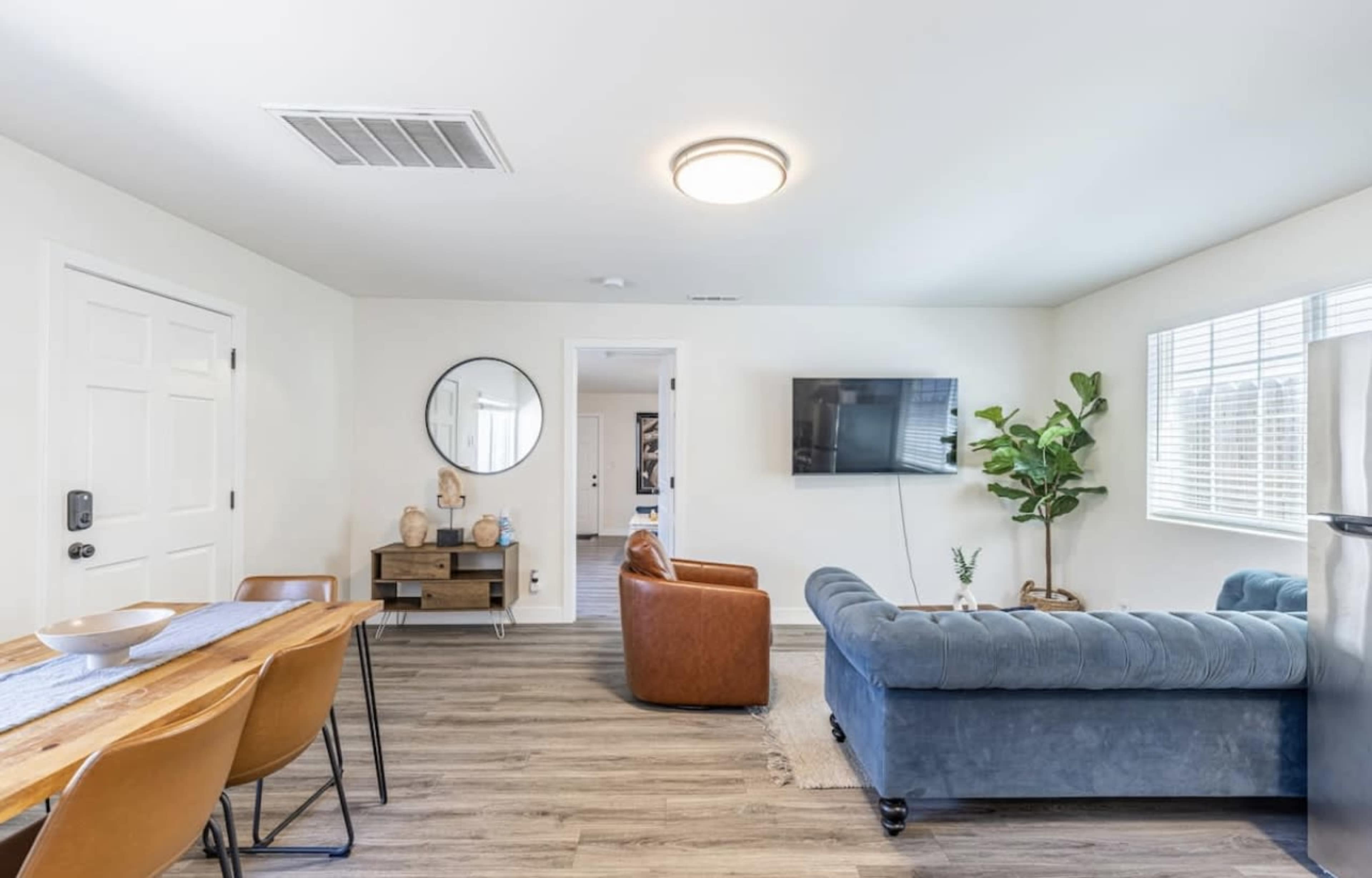 A modern living room and dining area with a blue couch, an armchair, a dining table, and a flat-screen TV mounted on the wall.