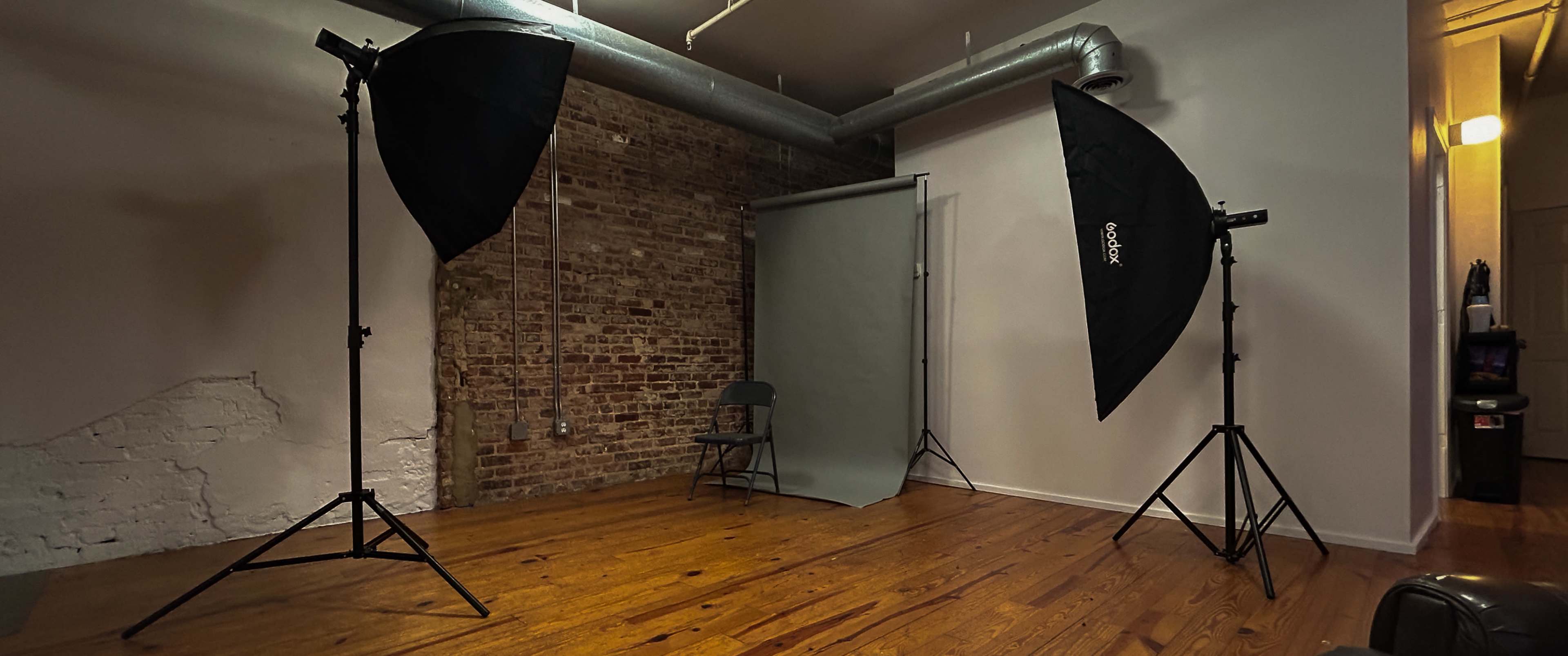 A photography studio with two large softbox lights, a backdrop stand with a gray backdrop, and a folding chair on a wooden floor.