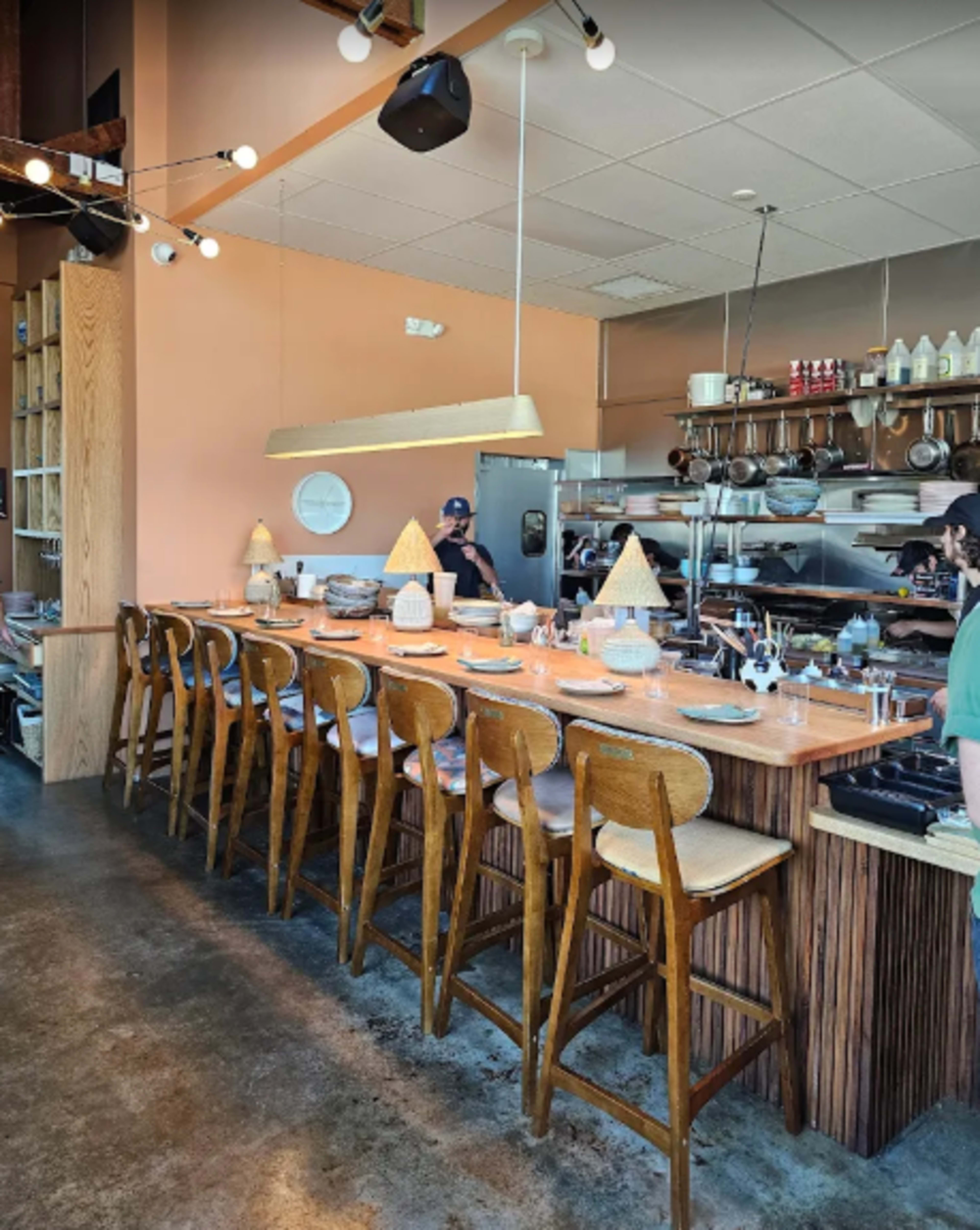 The image shows a modern restaurant interior with a long wooden bar, several bar stools, and an open kitchen area in the background.