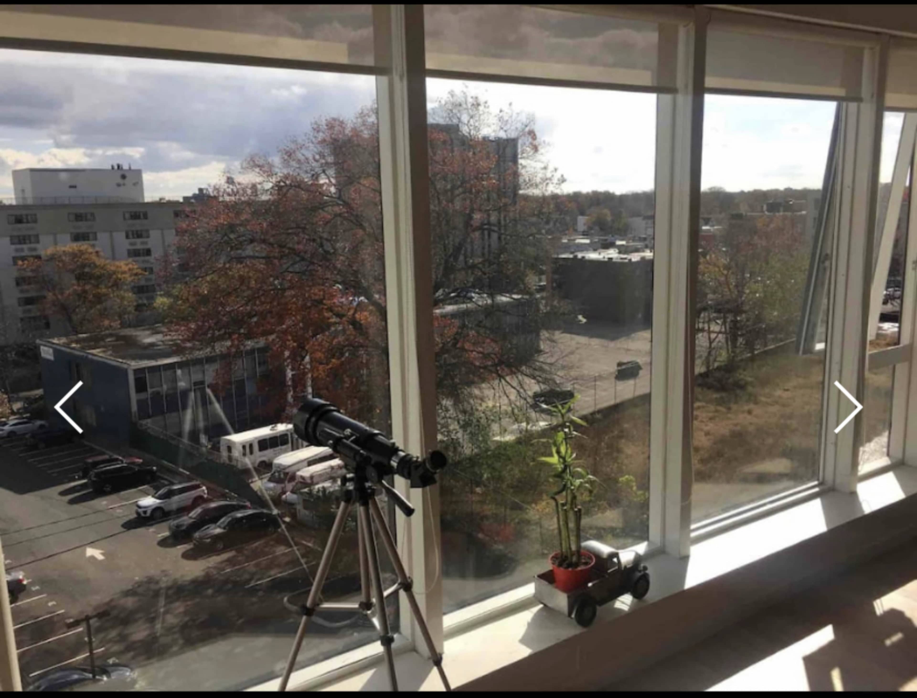 The image shows a large window view overlooking a parking lot and buildings, with a telescope and a potted plant on a windowsill inside.