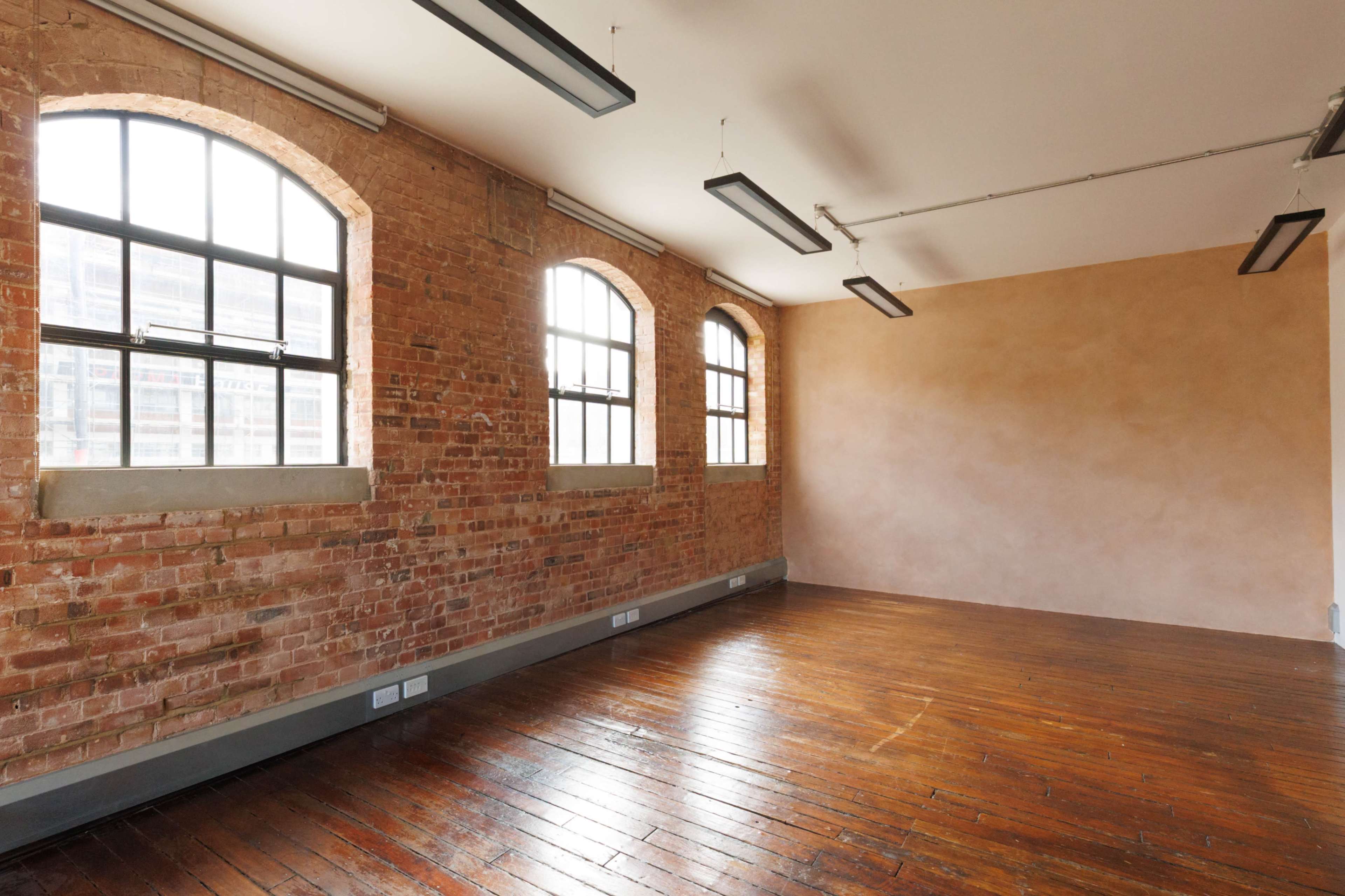 The image shows an empty room with exposed brick walls, large arched windows, and polished wooden floors.