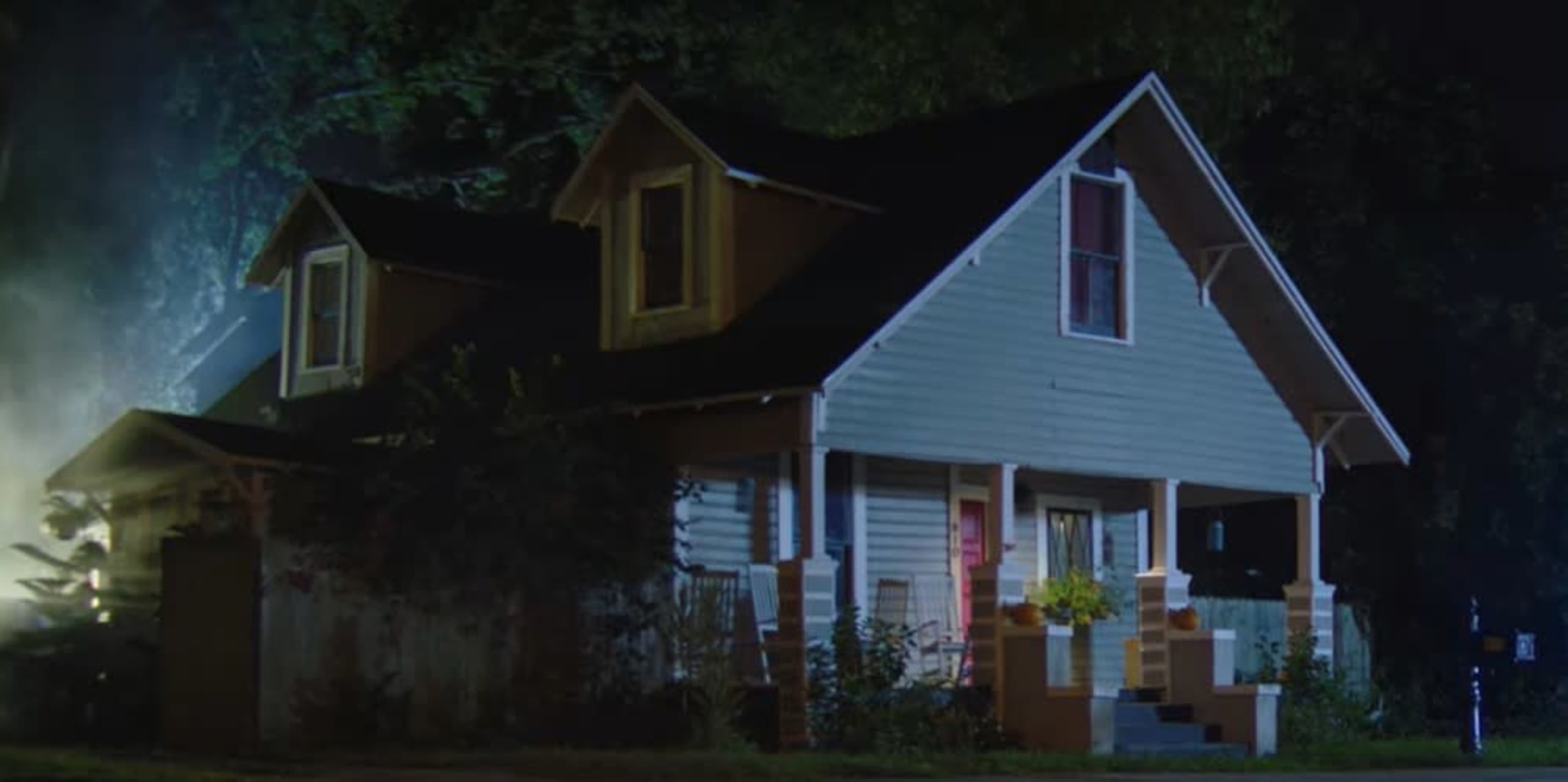 A two-story house with a sloped roof and a front porch is illuminated by a faint light in a dimly lit setting.