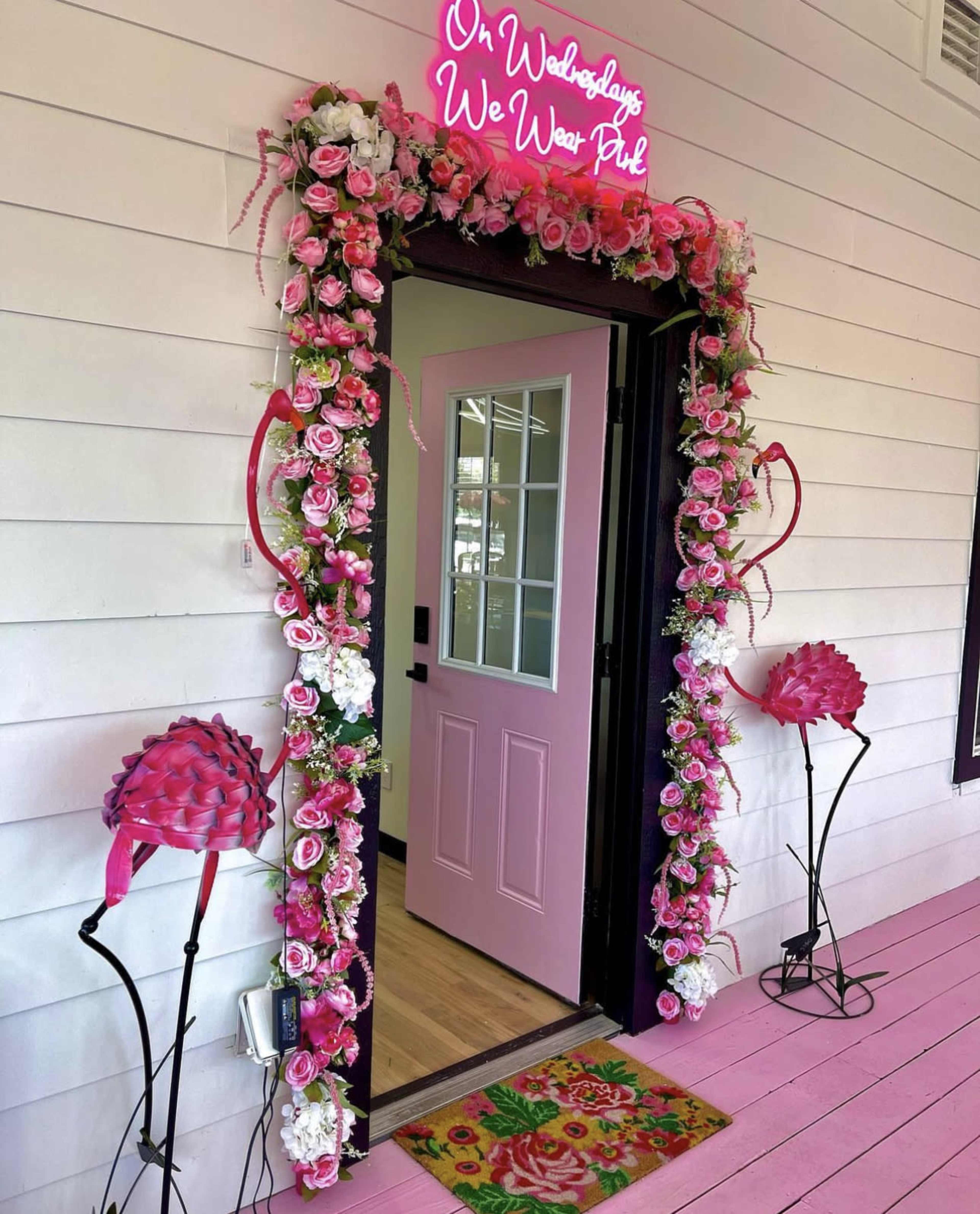 A brightly decorated entrance with a pink door, surrounded by a floral arch and a neon sign that reads "On Wednesdays We Wear Pink."