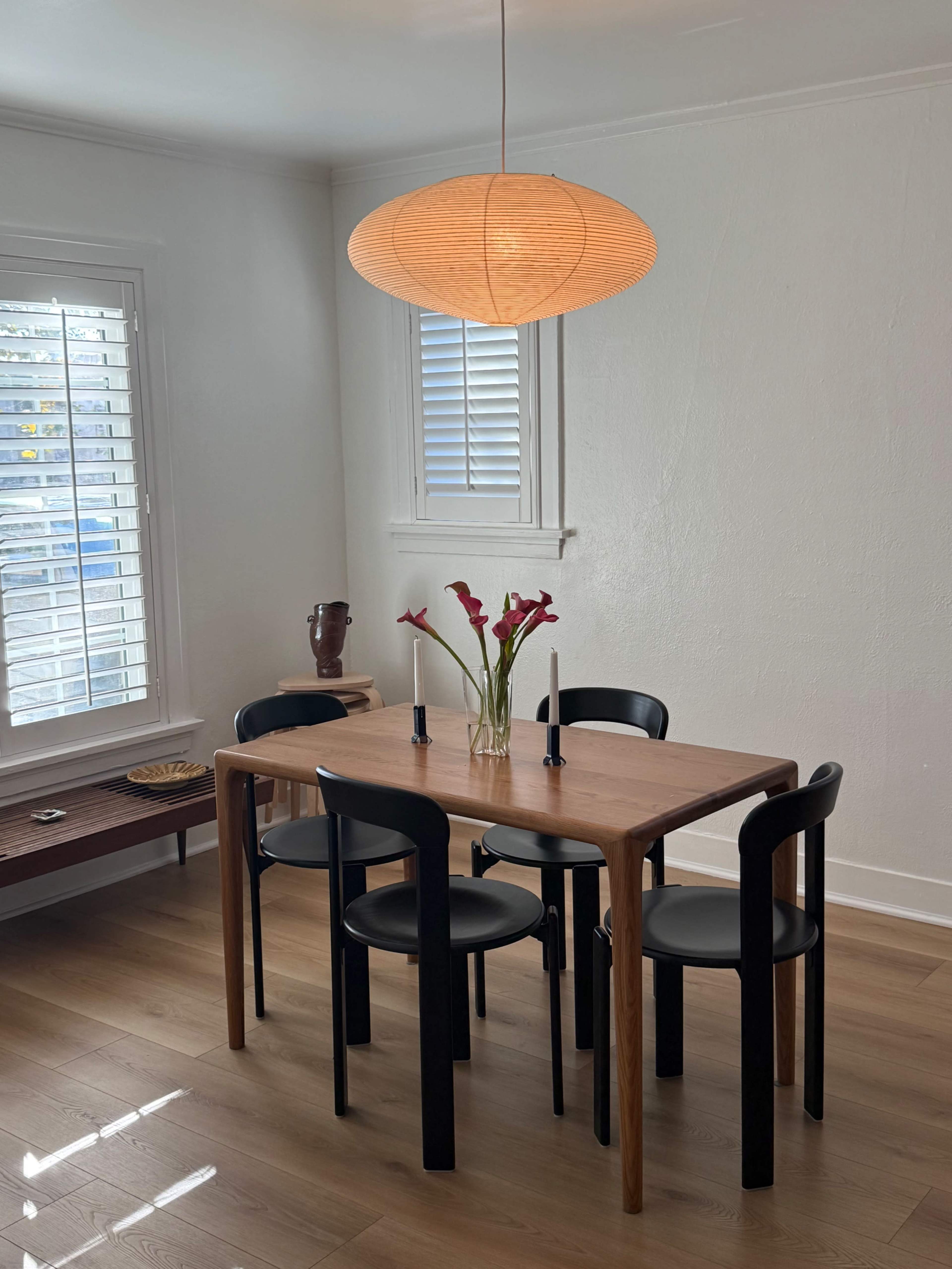 A wooden dining table with four black chairs is set in a bright room, featuring a hanging lantern light and a vase with flowers on the table.