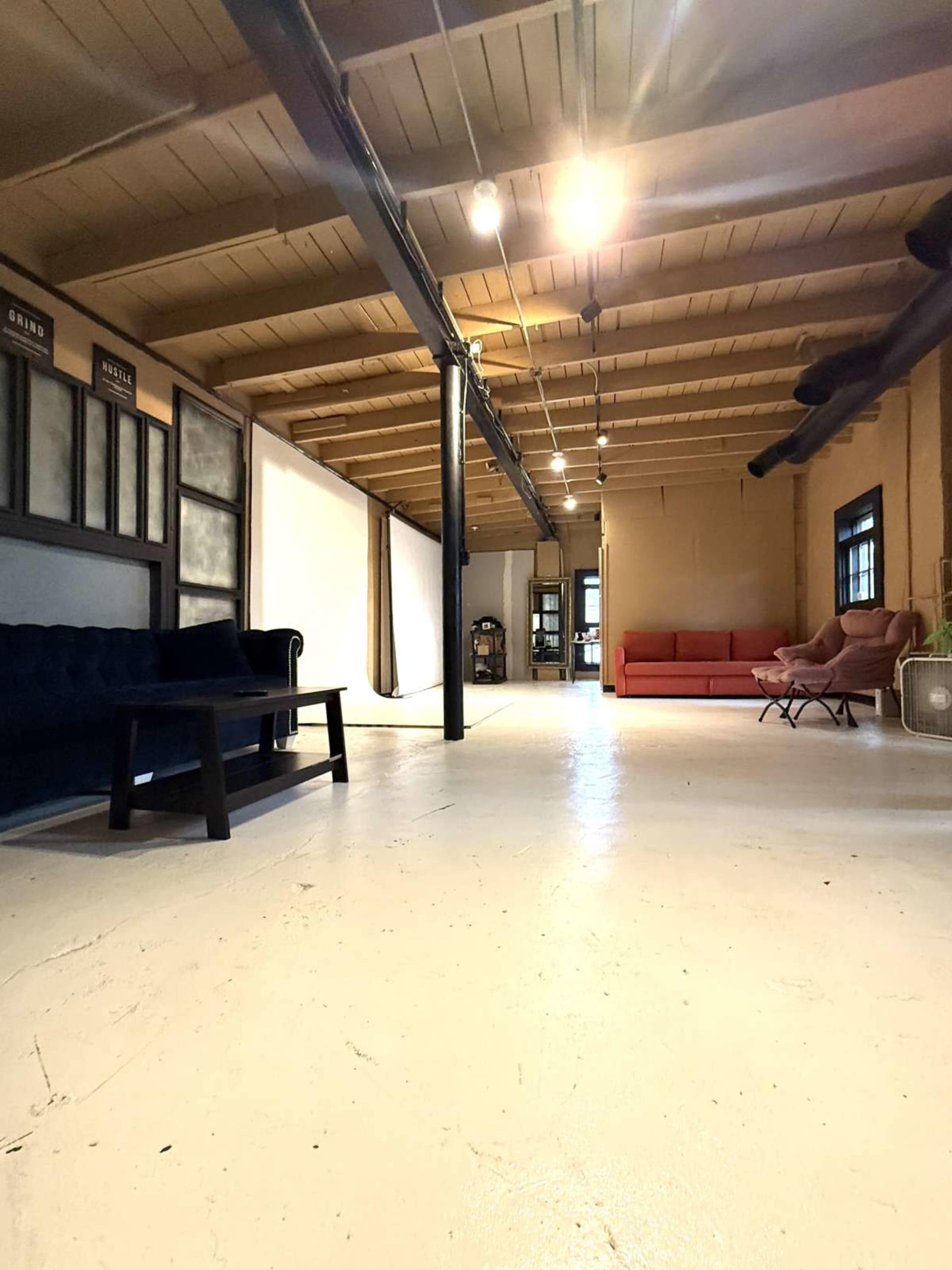 The image shows a spacious room with wooden beams, a polished white floor, and several seating areas including a black sofa and a red couch.