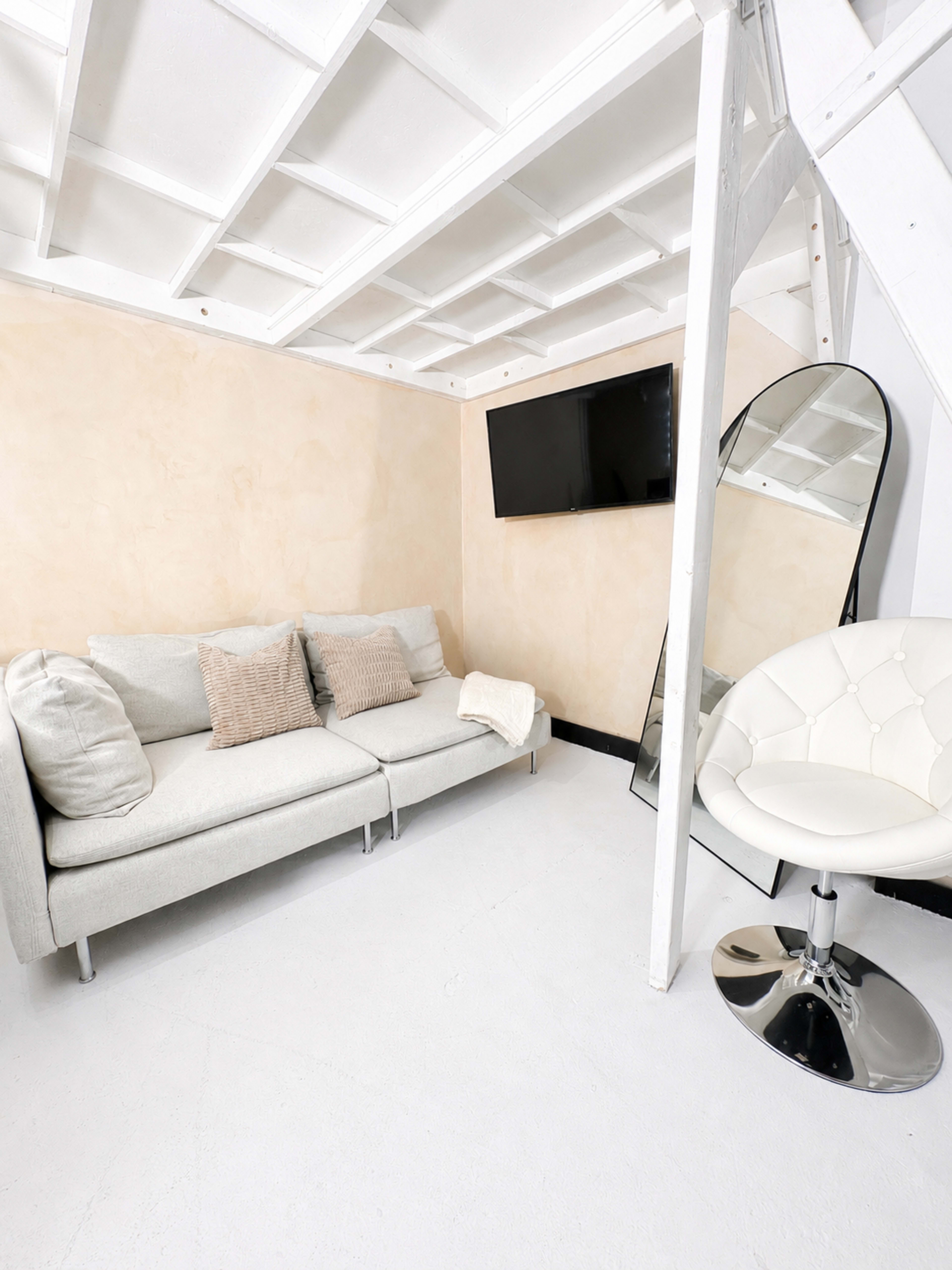 The image shows a small, minimalist living space featuring a light gray sofa with decorative pillows, a flat-screen TV mounted on the wall, a large mirror, and a modern white chair.