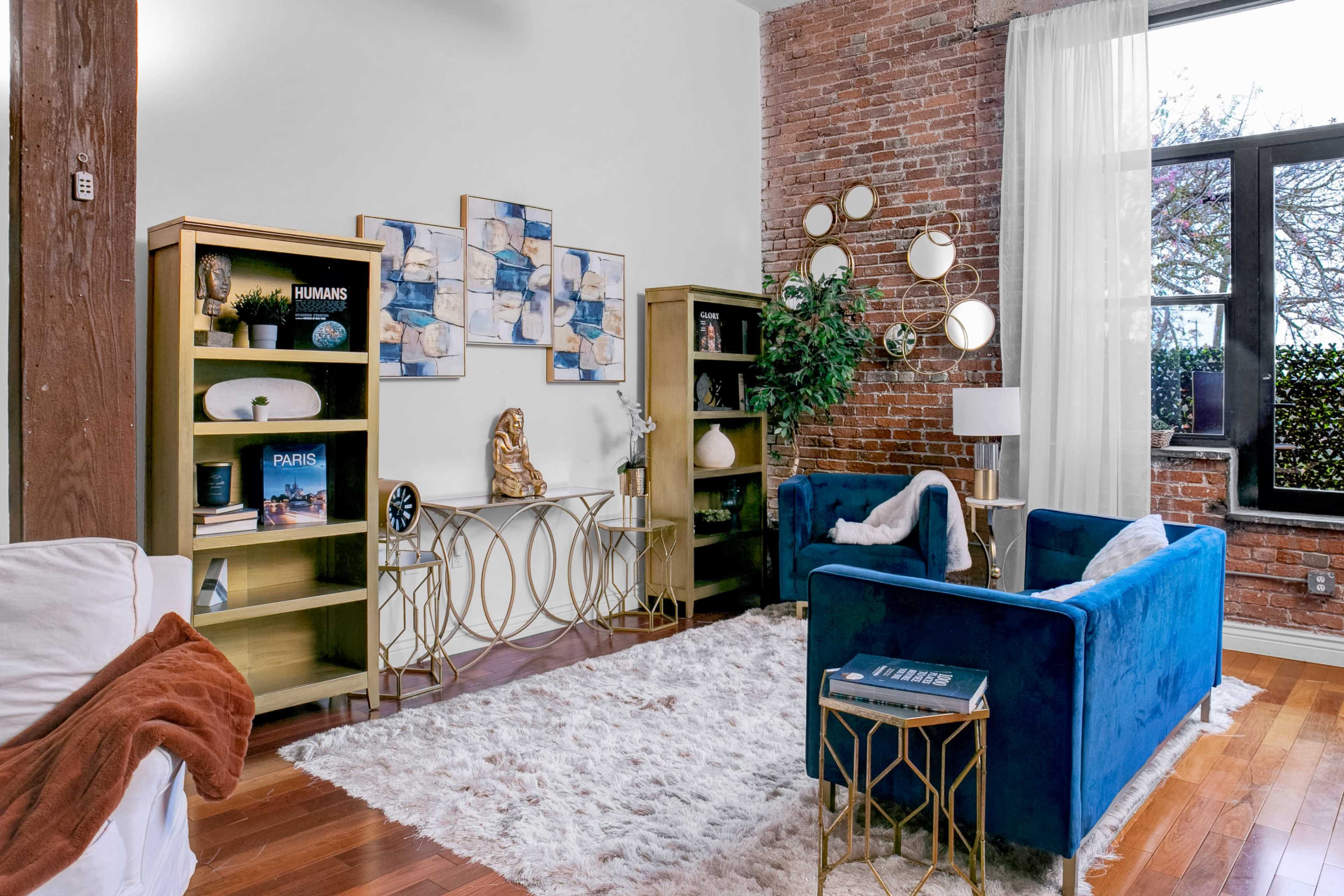 The image shows a modern living room with exposed brick walls, featuring blue sofas, gold-accented furniture, and decorative artwork.