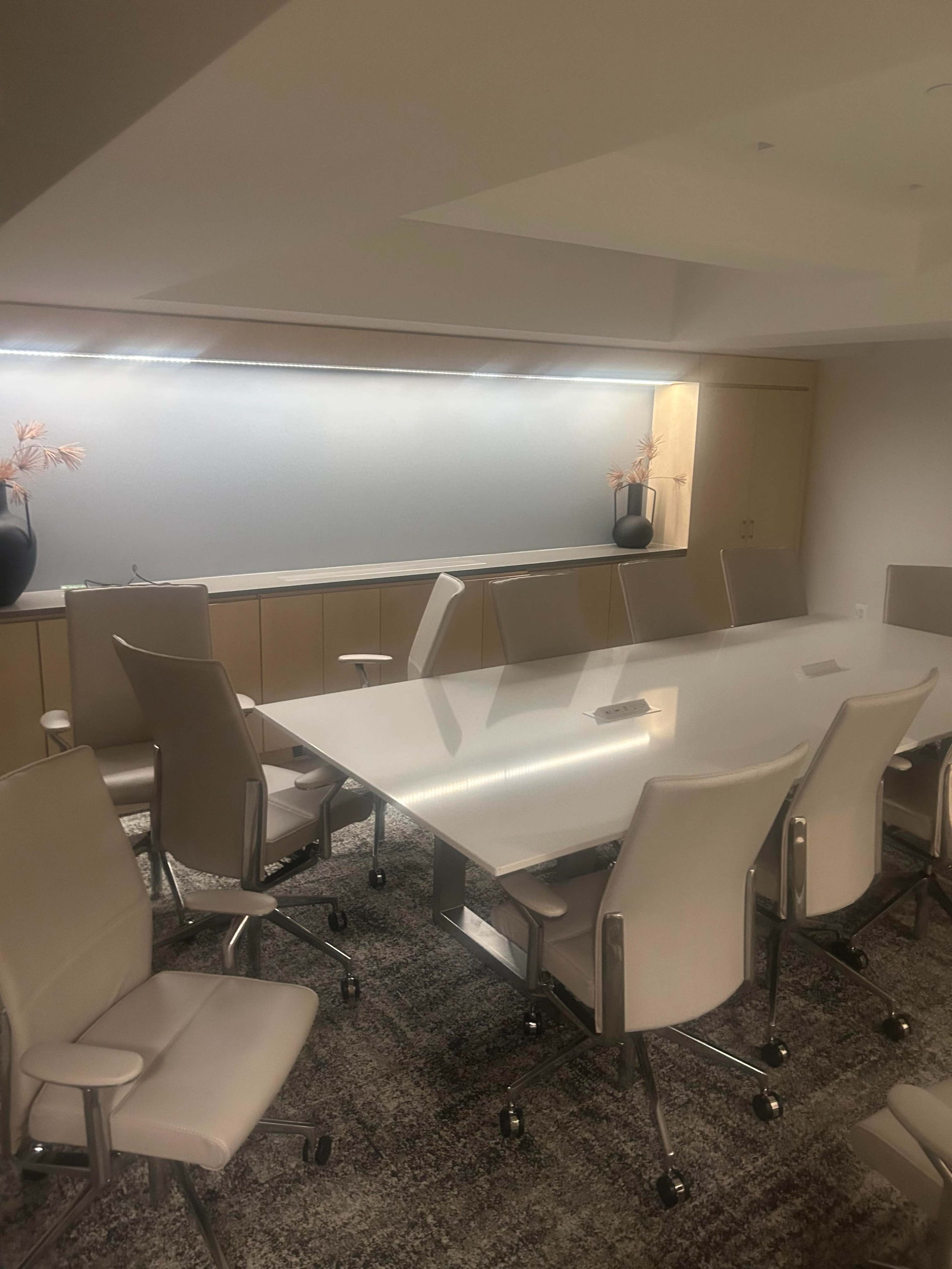 A modern conference room features a long white table surrounded by several beige chairs and a neutral-colored carpet, with a cabinet and plants in the background.