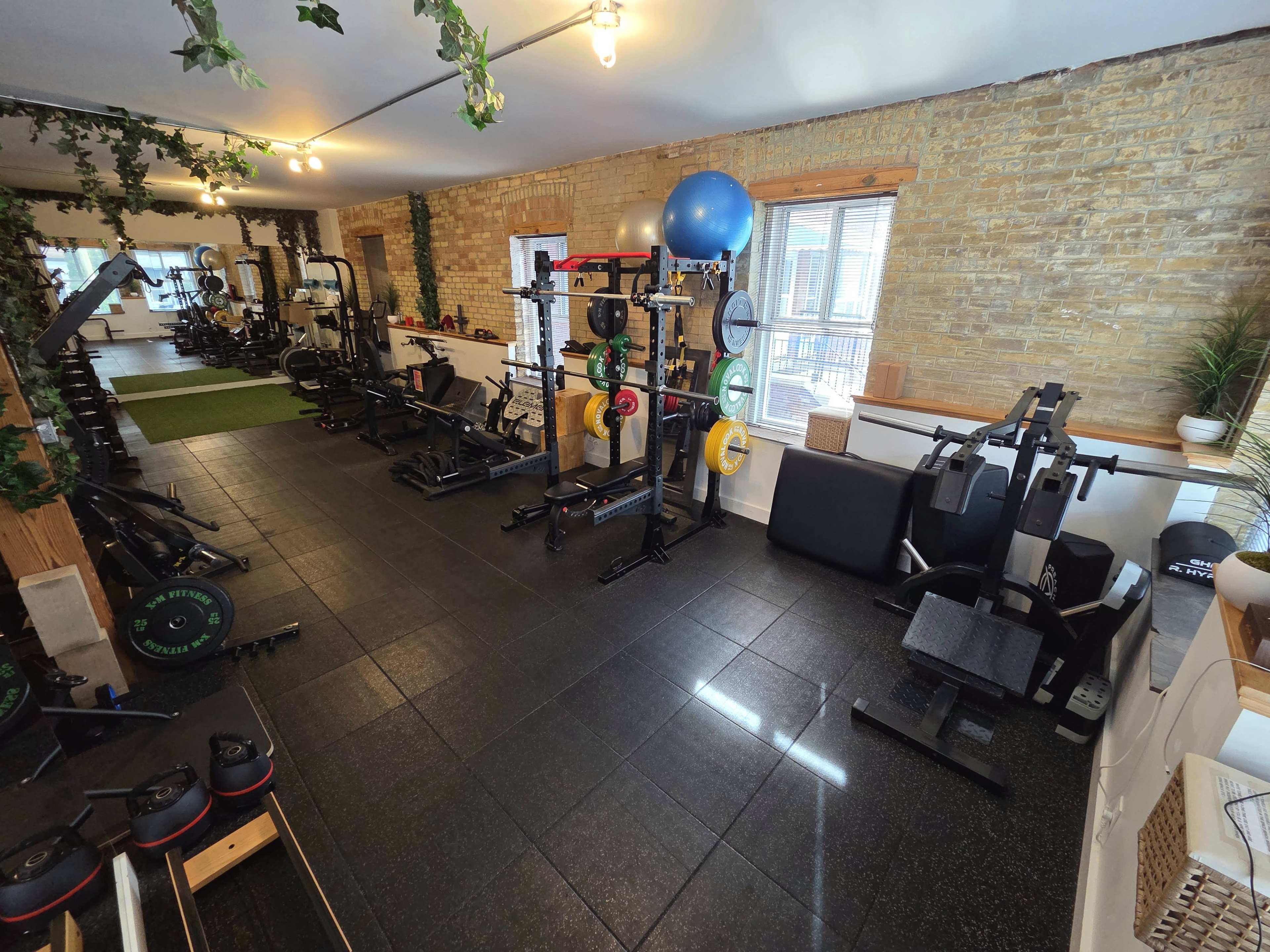The image shows a well-equipped gym with various weightlifting and exercise machines arranged on a rubber flooring surface.