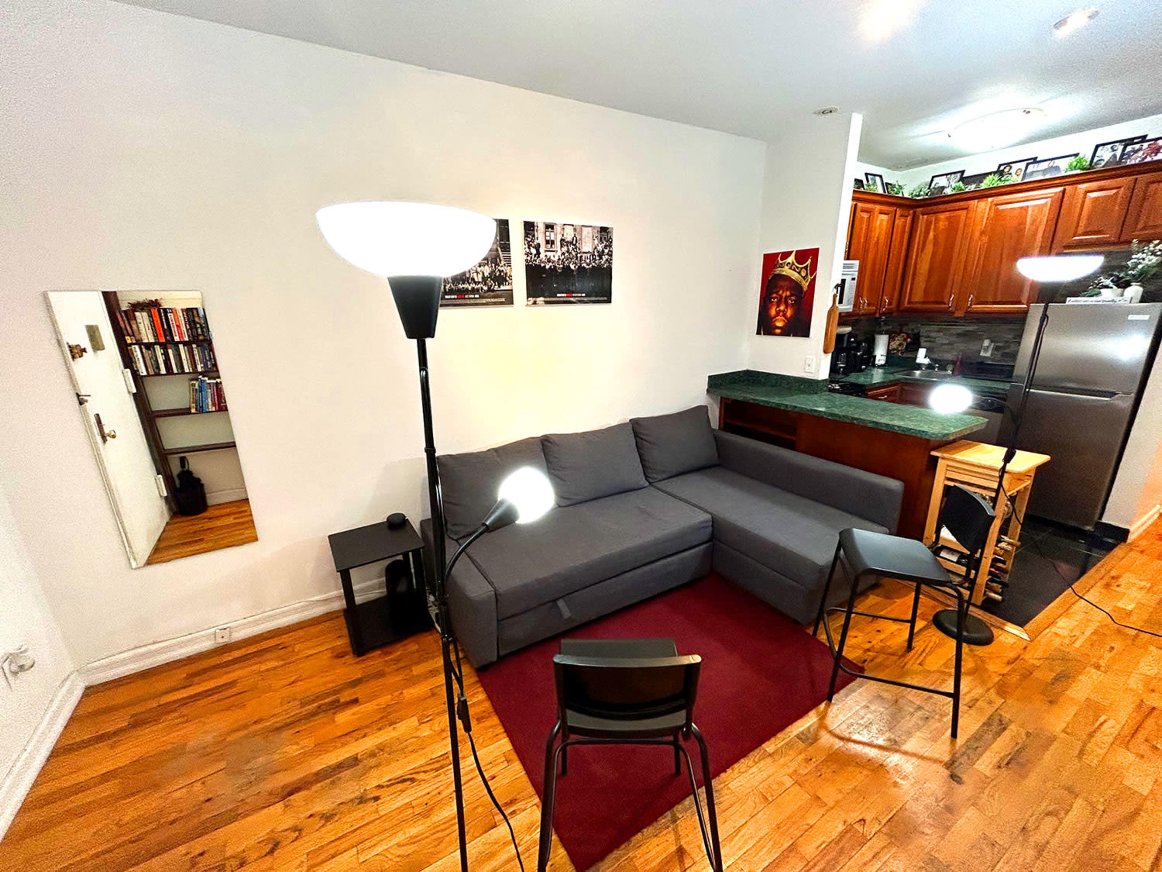 A compact living room with a gray sectional sofa, two floor lamps, a small round table, and a kitchen area in the background.