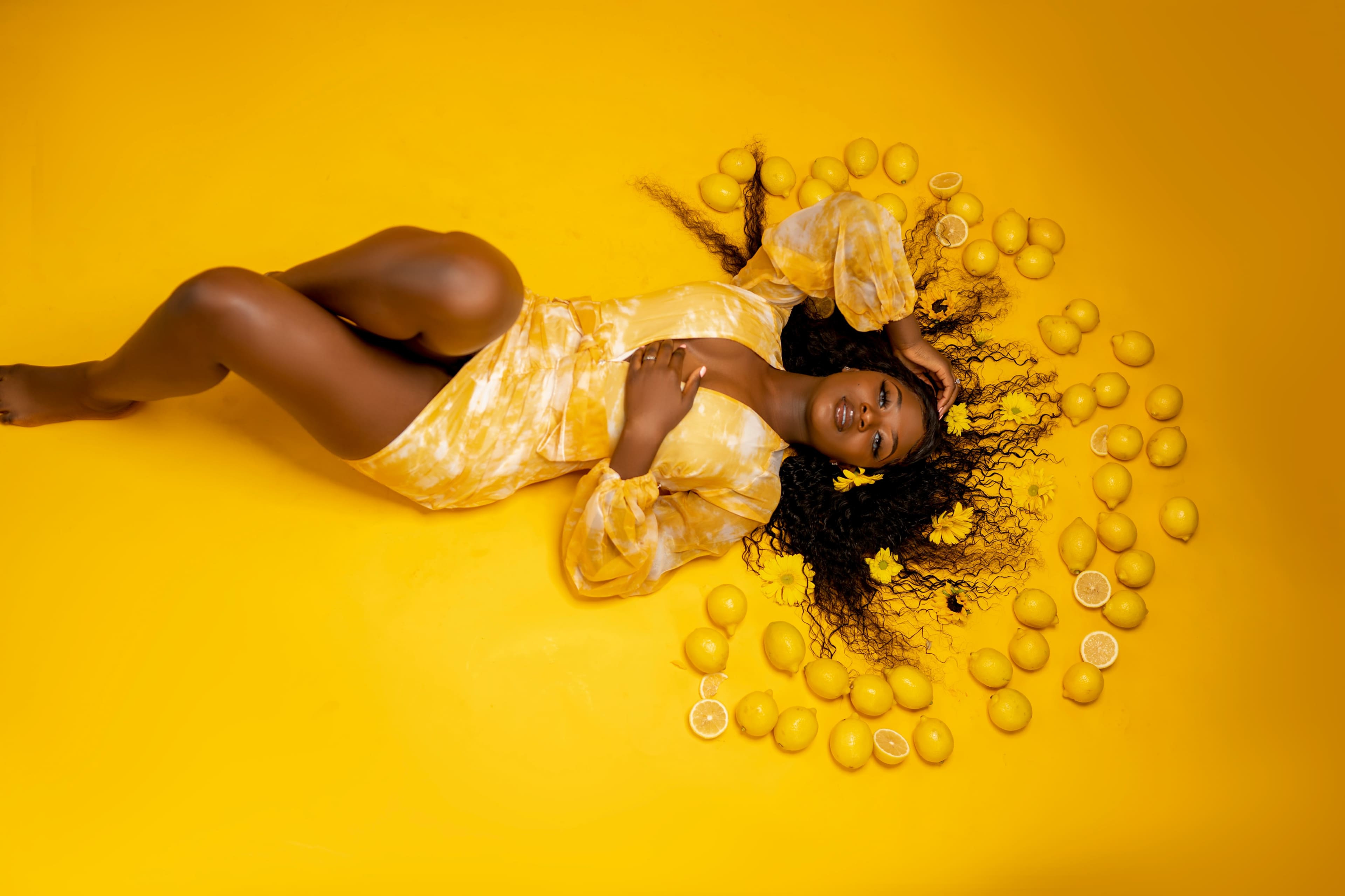 A person in a yellow outfit lies on a yellow floor surrounded by lemons and flower petals.