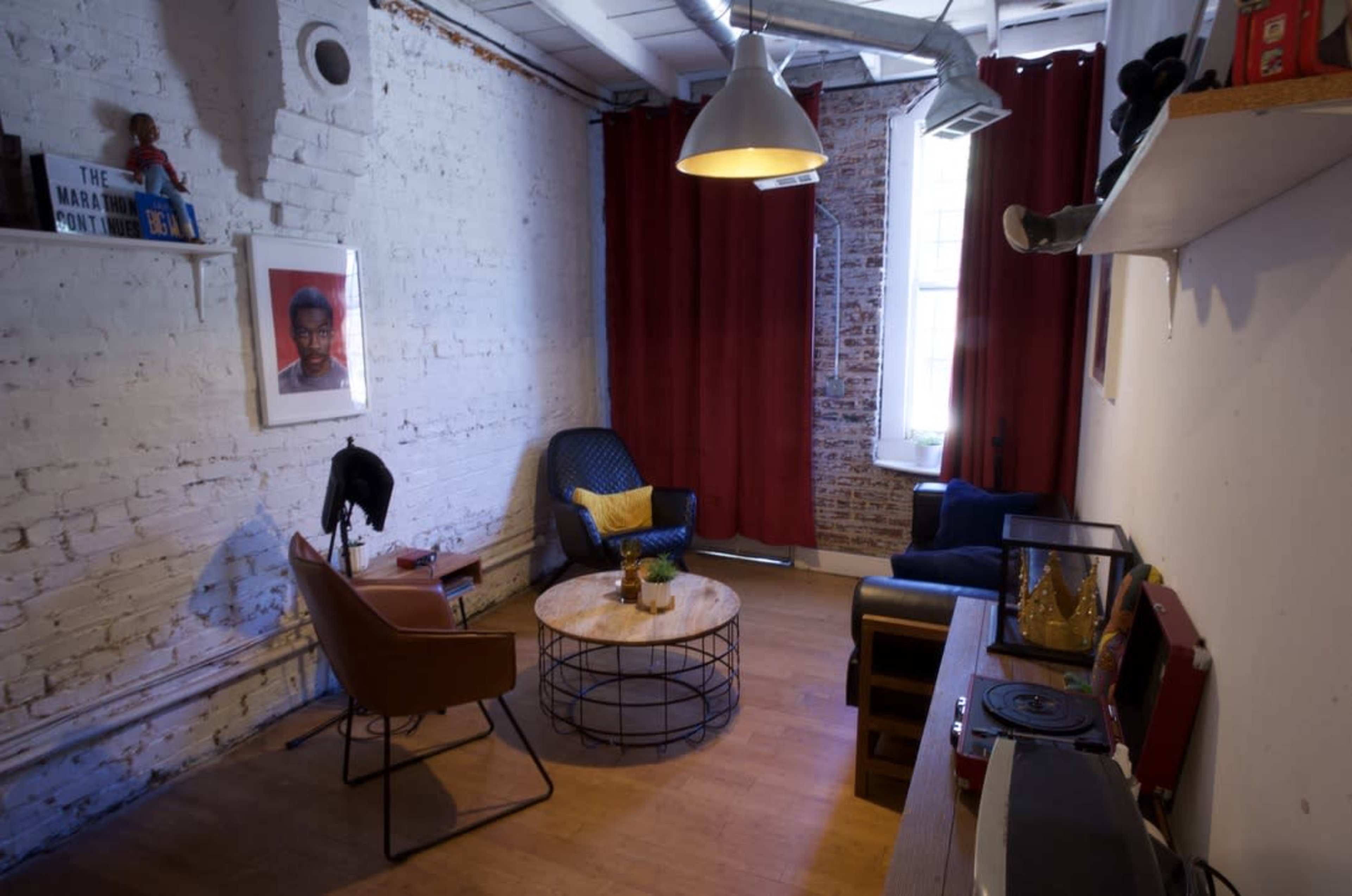 A cozy room with exposed brick walls, two armchairs, a round coffee table, and large red curtains covering a window.