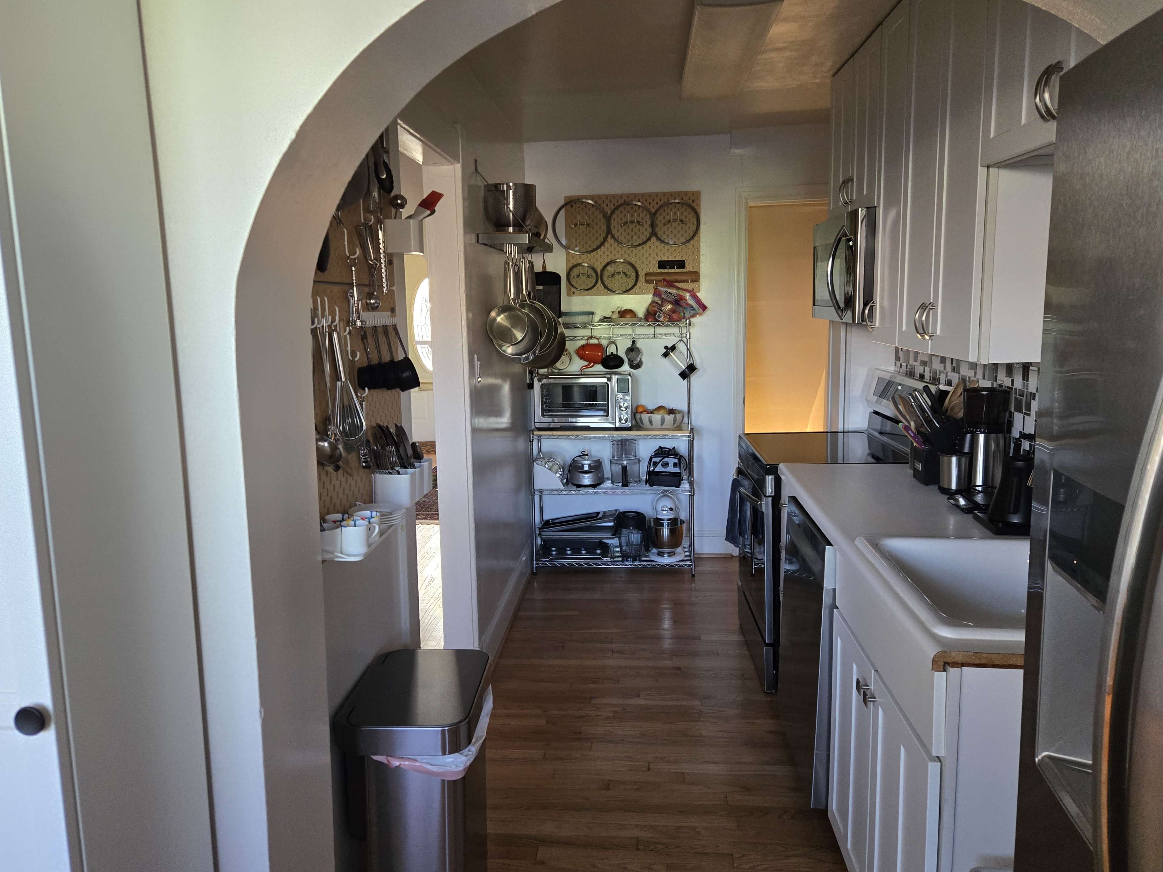 The image shows a narrow kitchen with white cabinetry, a stainless steel refrigerator, and various kitchen appliances arranged along the walls.