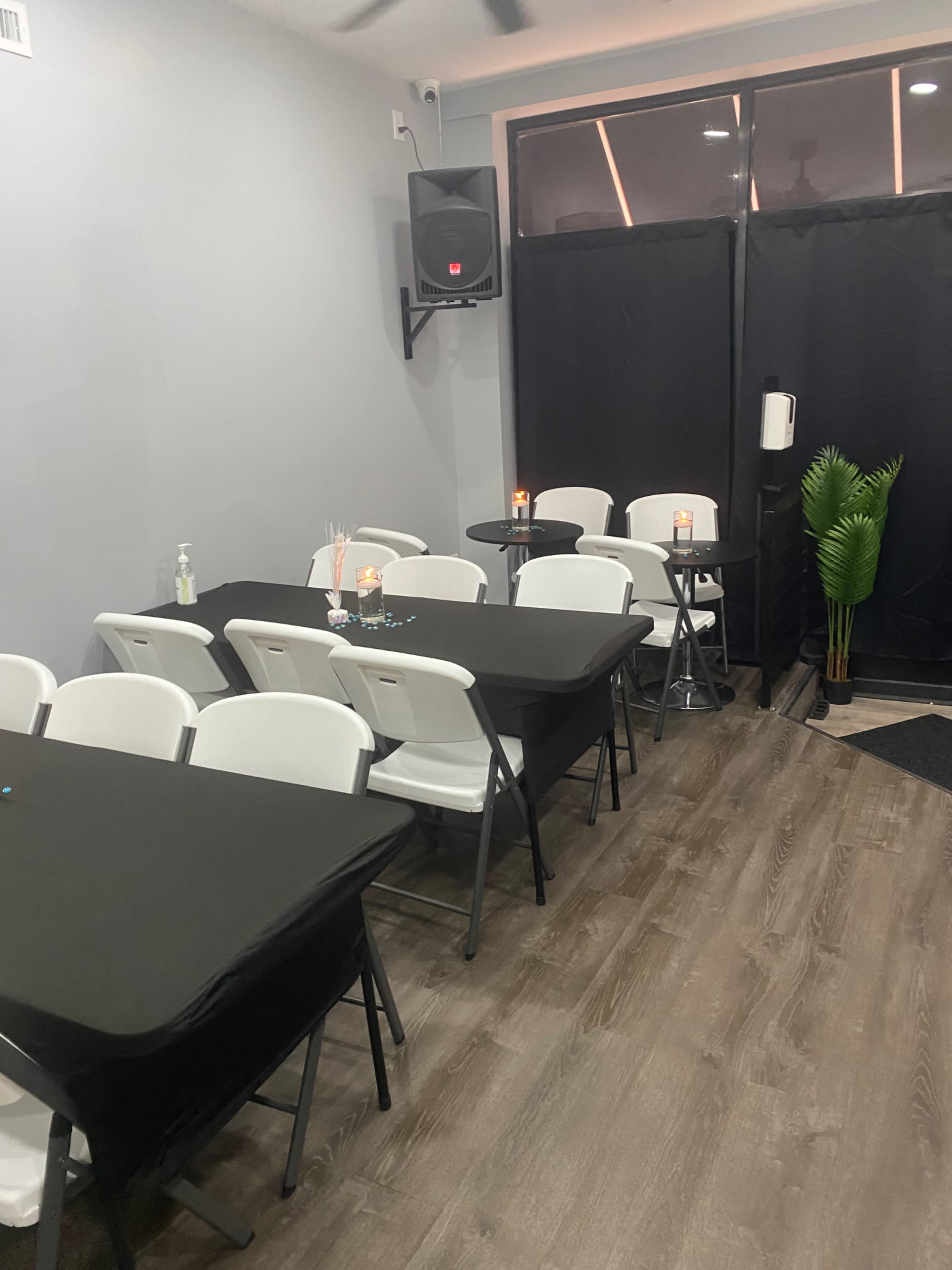 The image shows a modern, sparsely furnished room with several black tables and white chairs arranged neatly, along with a small plant in the corner.