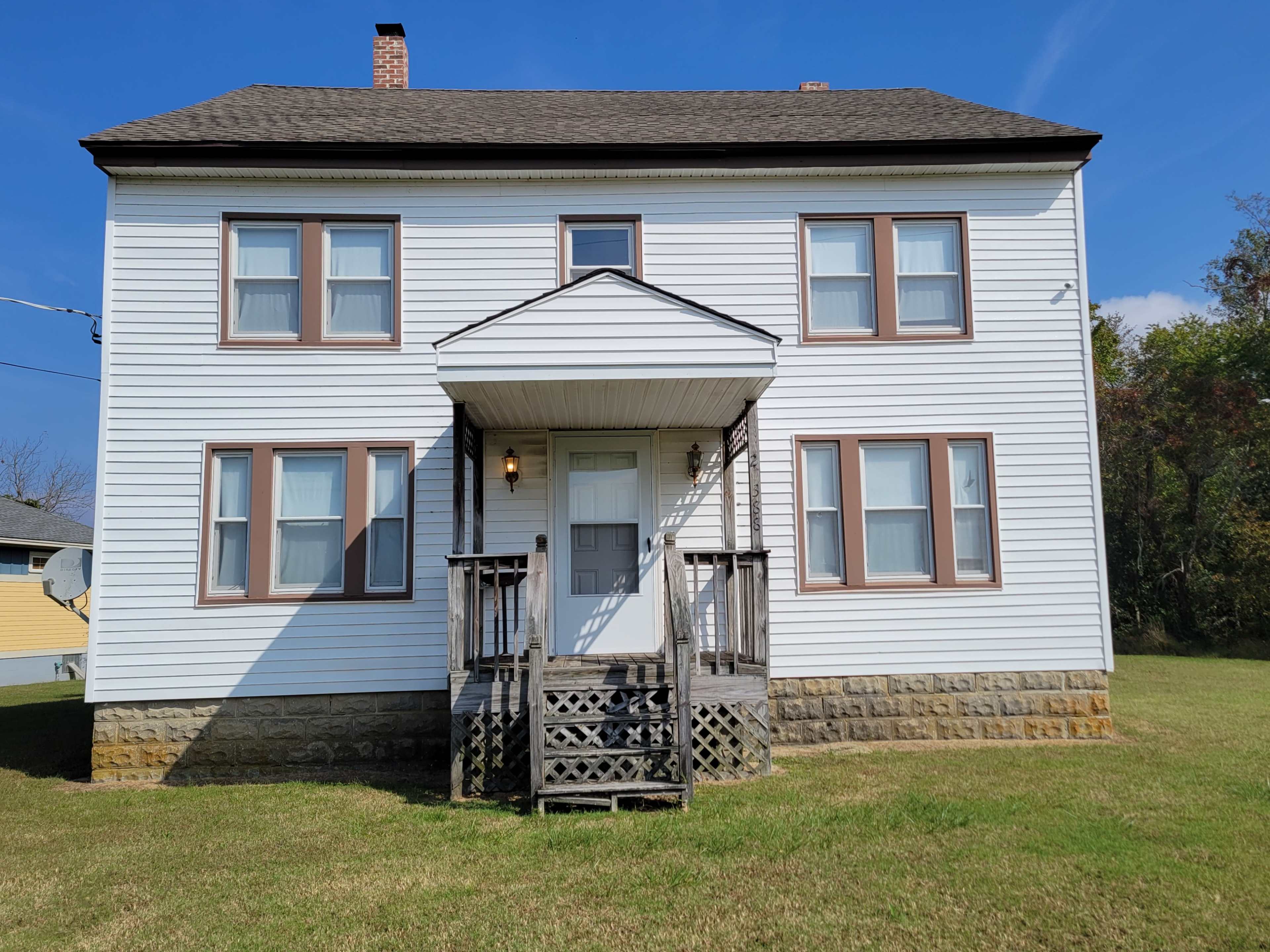 A two-story white house with a staircase and a small porch at the front, surrounded by a grassy yard.