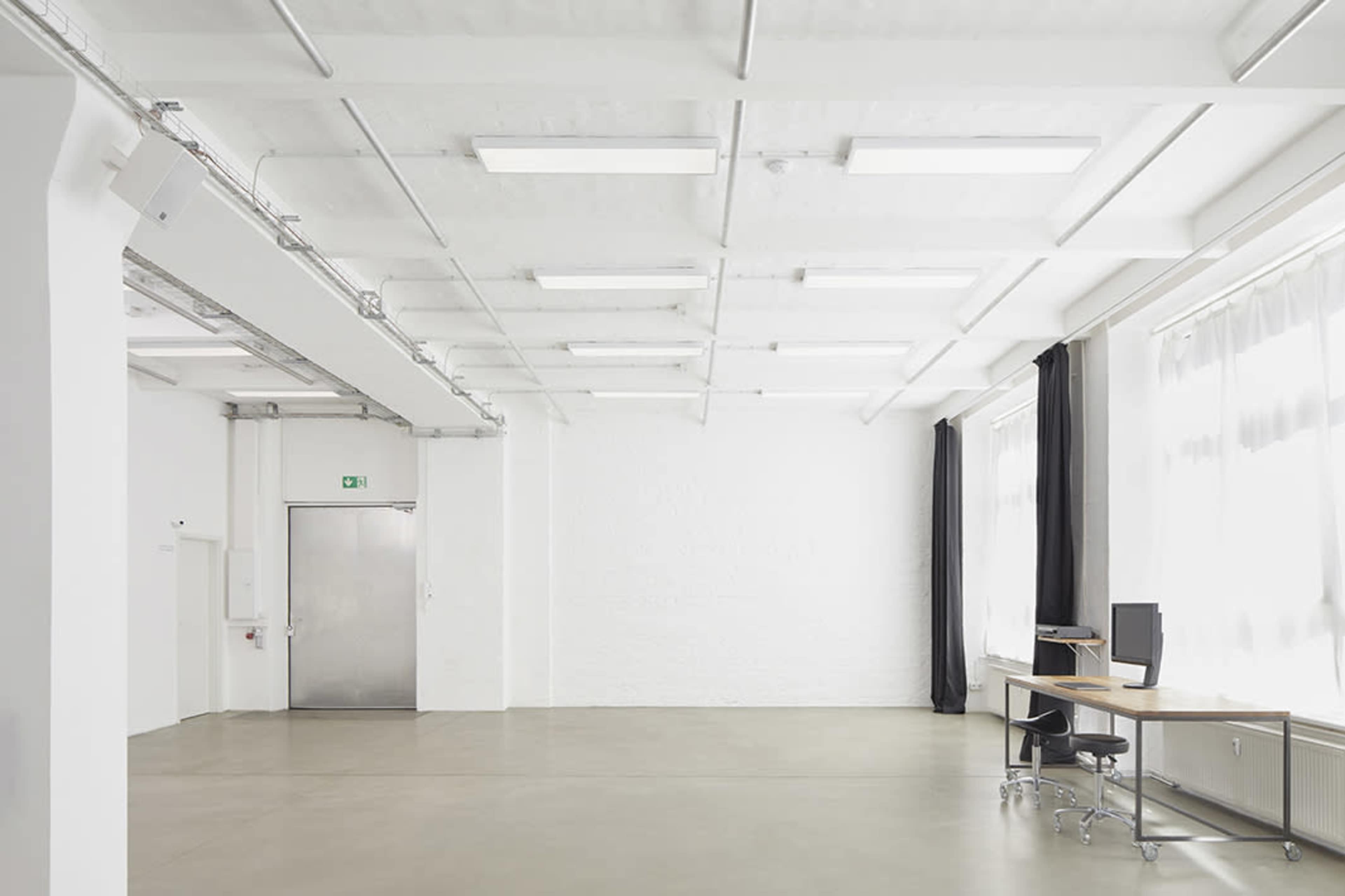 A bright, spacious office interior features a large window, a desk with a computer, and a metal door.