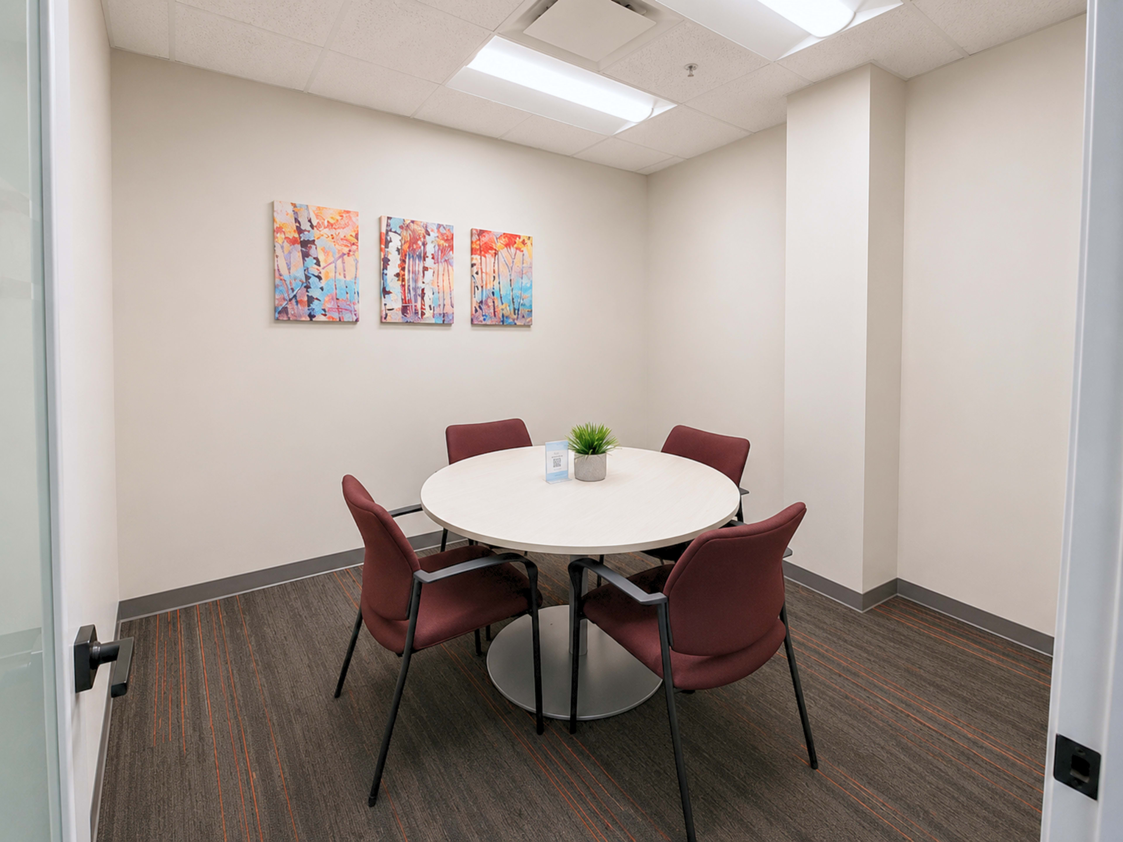 The image shows a small conference room with a round table surrounded by four maroon chairs and three colorful abstract paintings on the wall.
