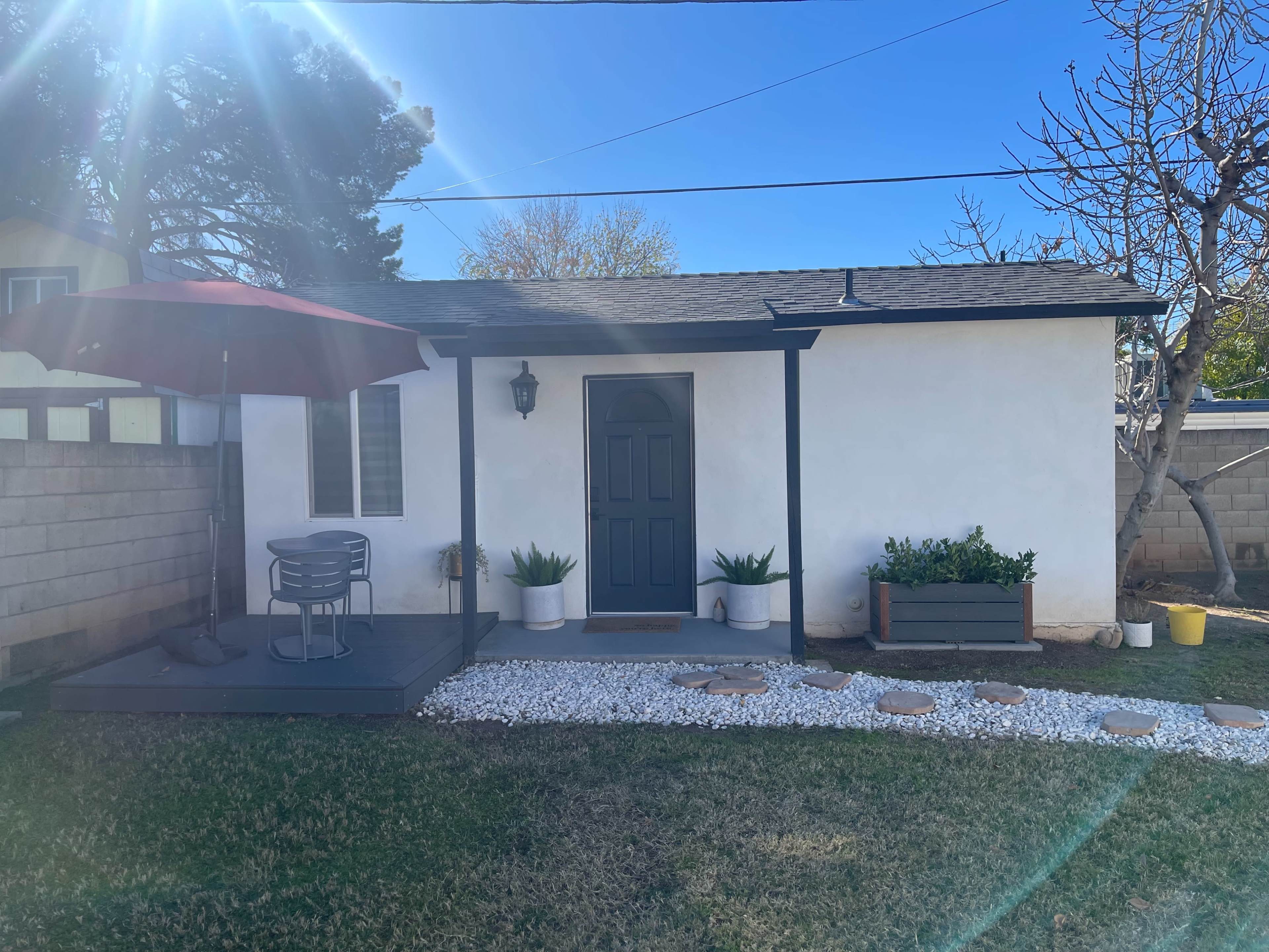 A small white cottage with a dark door and a porch area featuring a table and chairs is surrounded by gravel and grass.