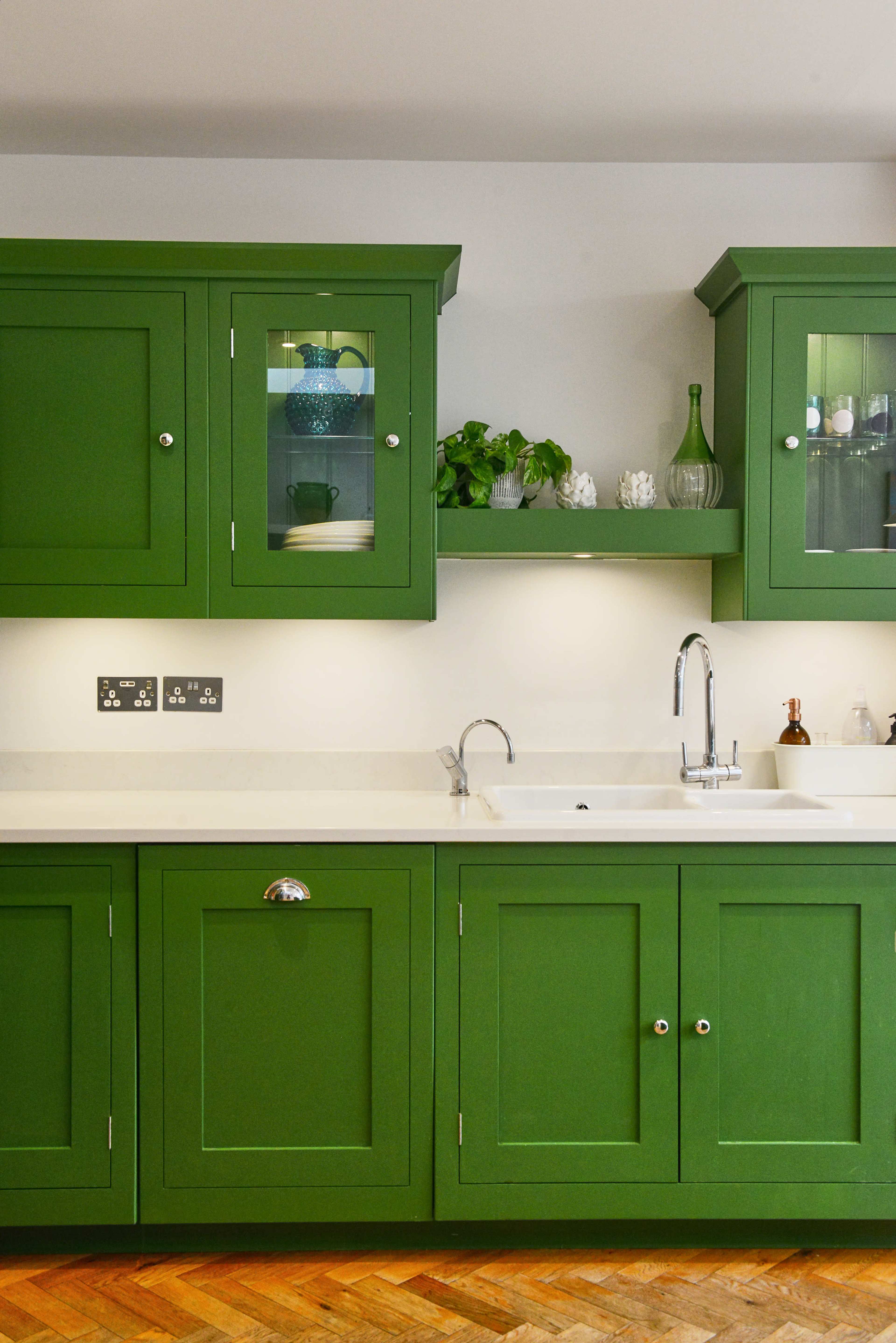 A kitchen with green cabinets, a white countertop, and a sink under a shelf containing decorative items.