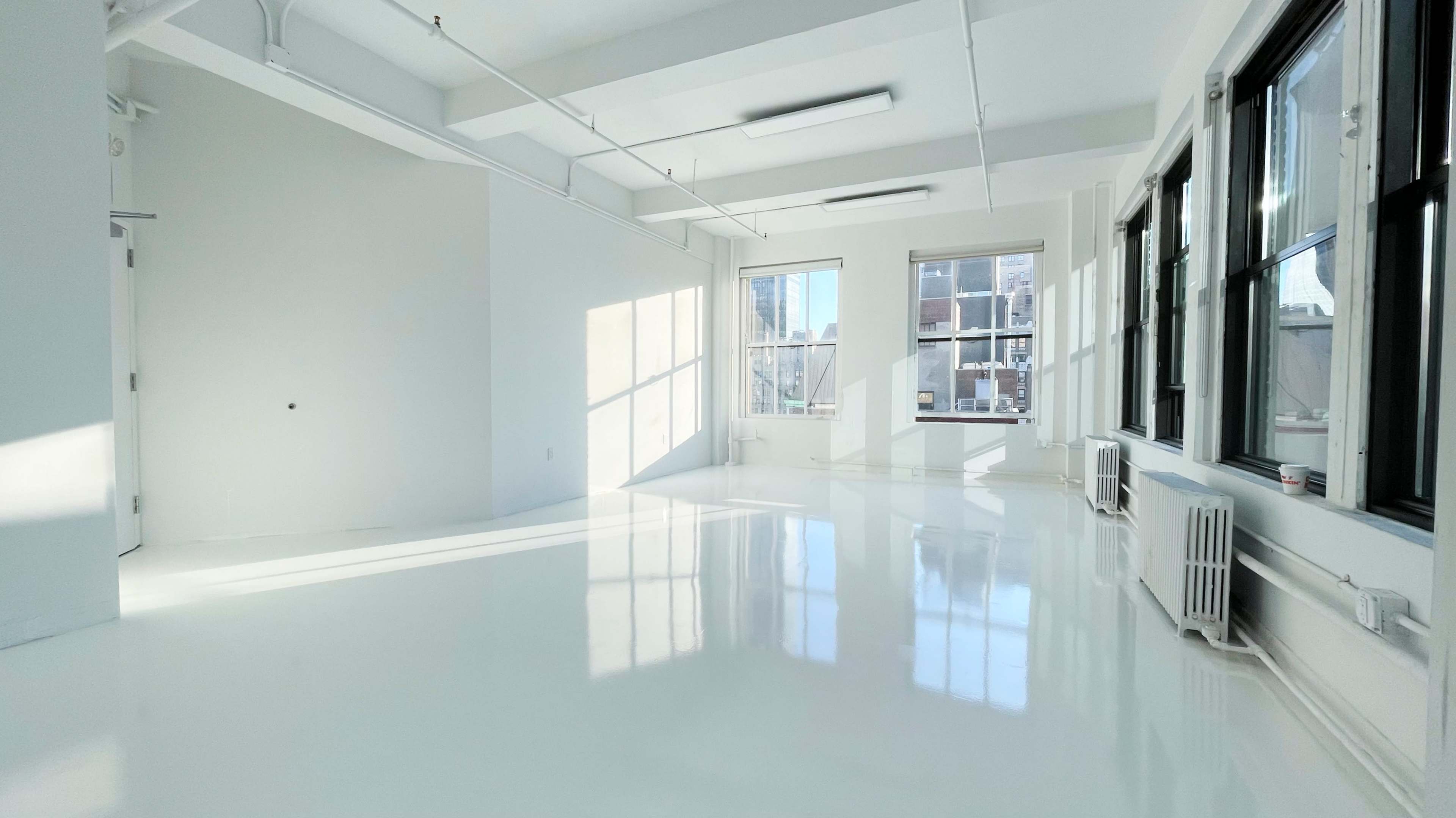 A spacious, brightly lit room with white walls and a glossy white floor, featuring large windows that let in natural light.