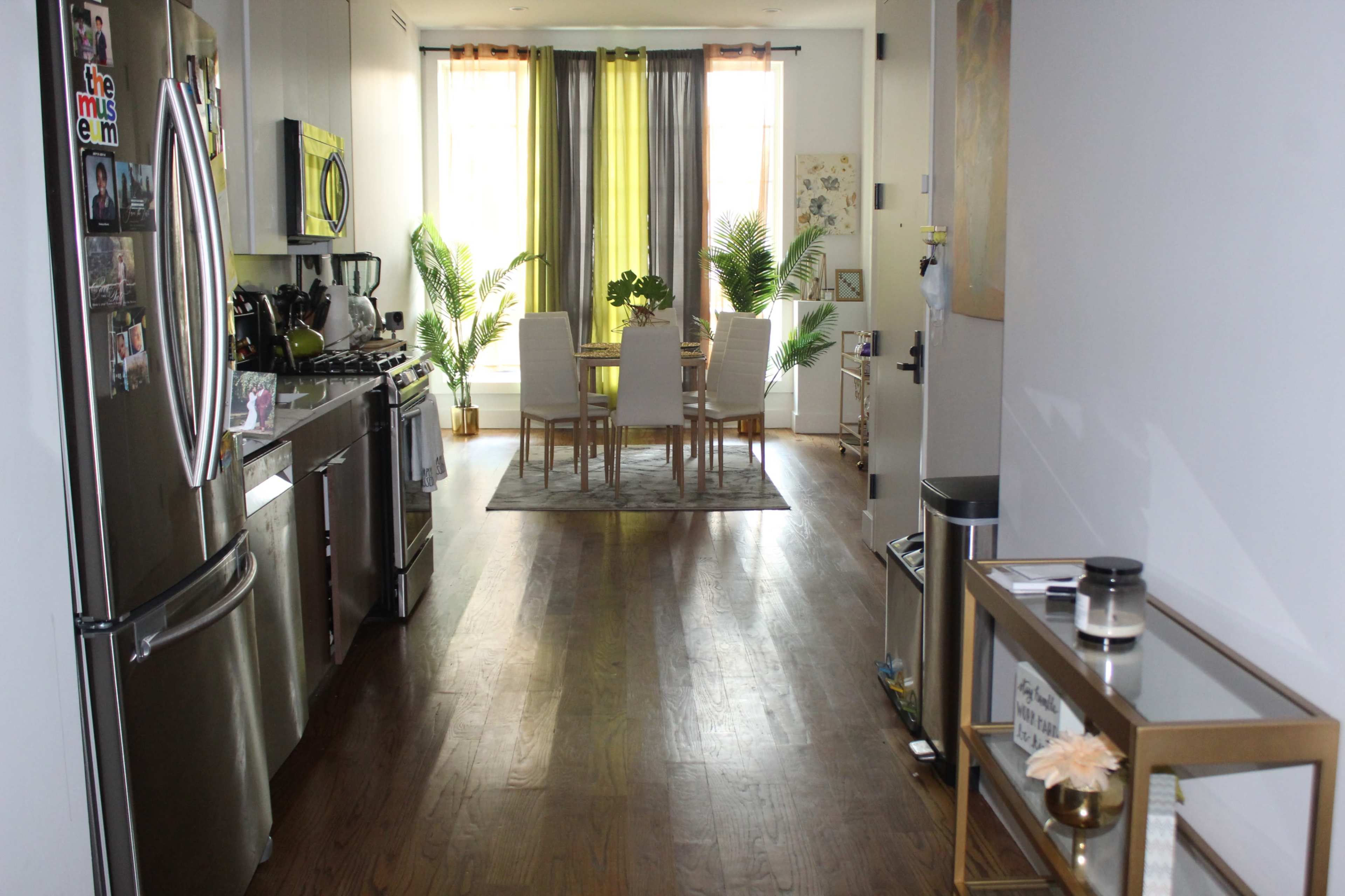 A spacious kitchen and dining area features a long wooden table surrounded by white chairs, with large windows covered by green curtains allowing natural light to fill the room.