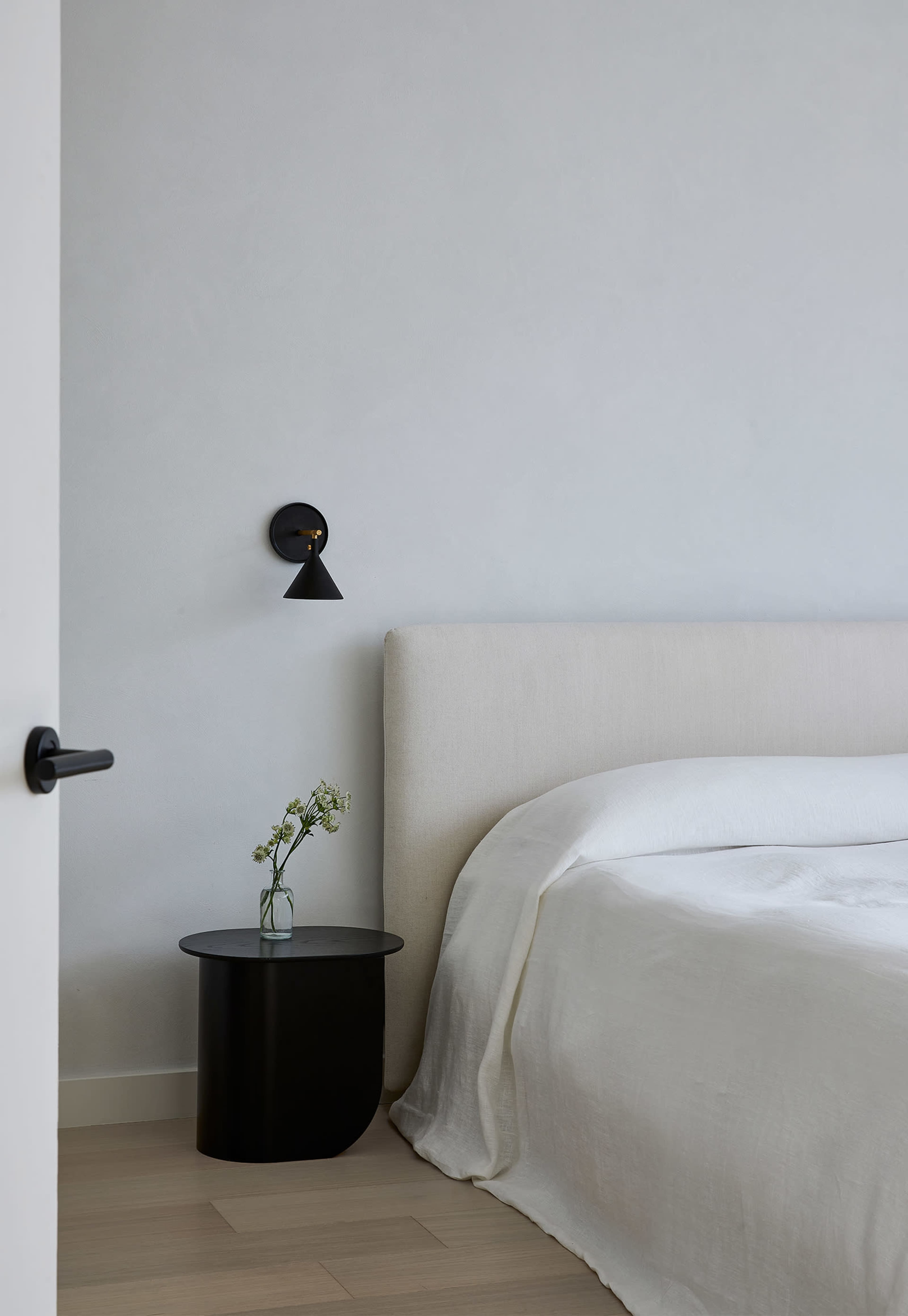 A minimalist bedroom features a light-colored bed with a plain duvet, a black side table holding a small vase of flowers, and a wall-mounted lamp.