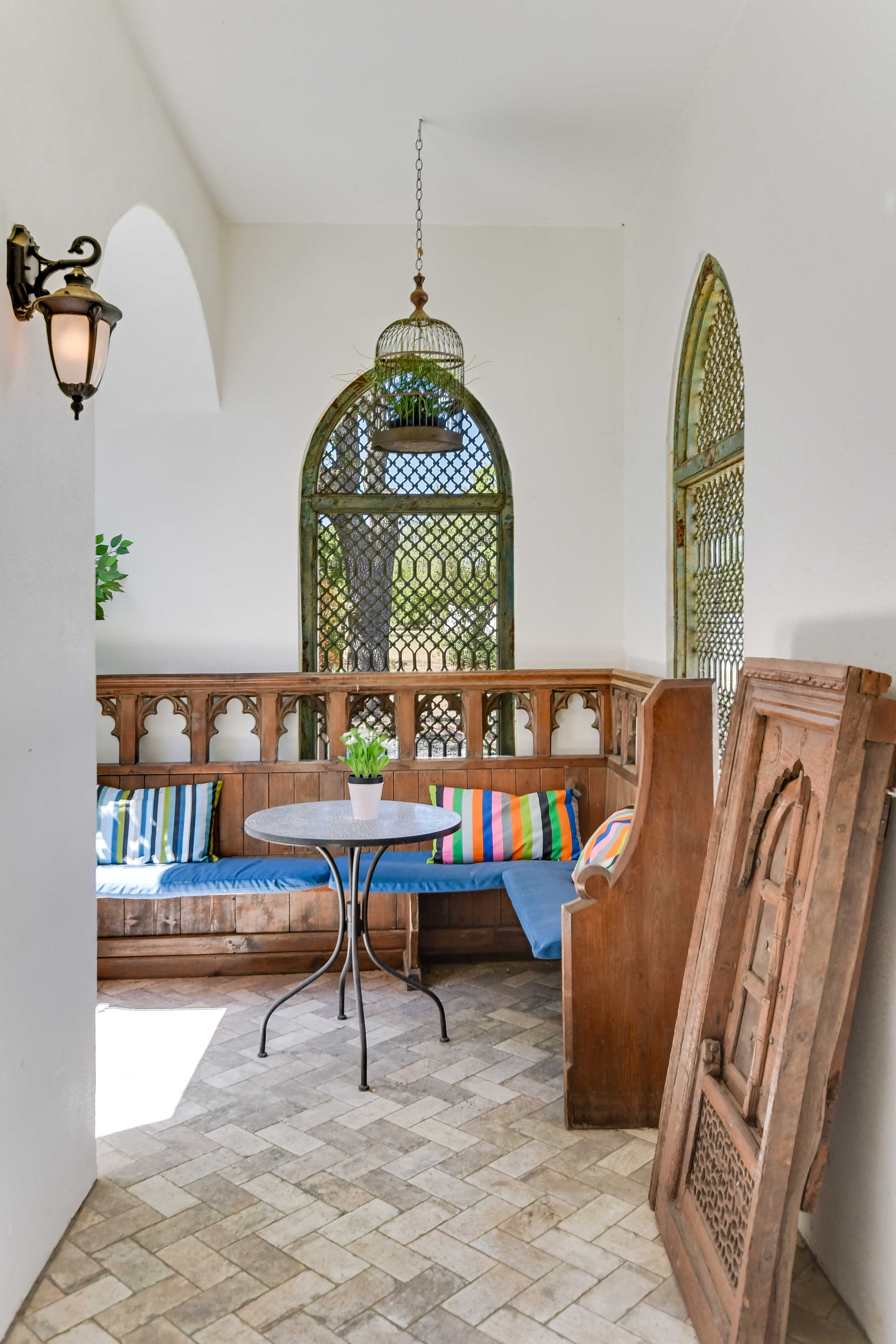 A small, bright sitting area features a wooden bench with colorful cushions, a round table, and an ornate wooden panel against the wall.