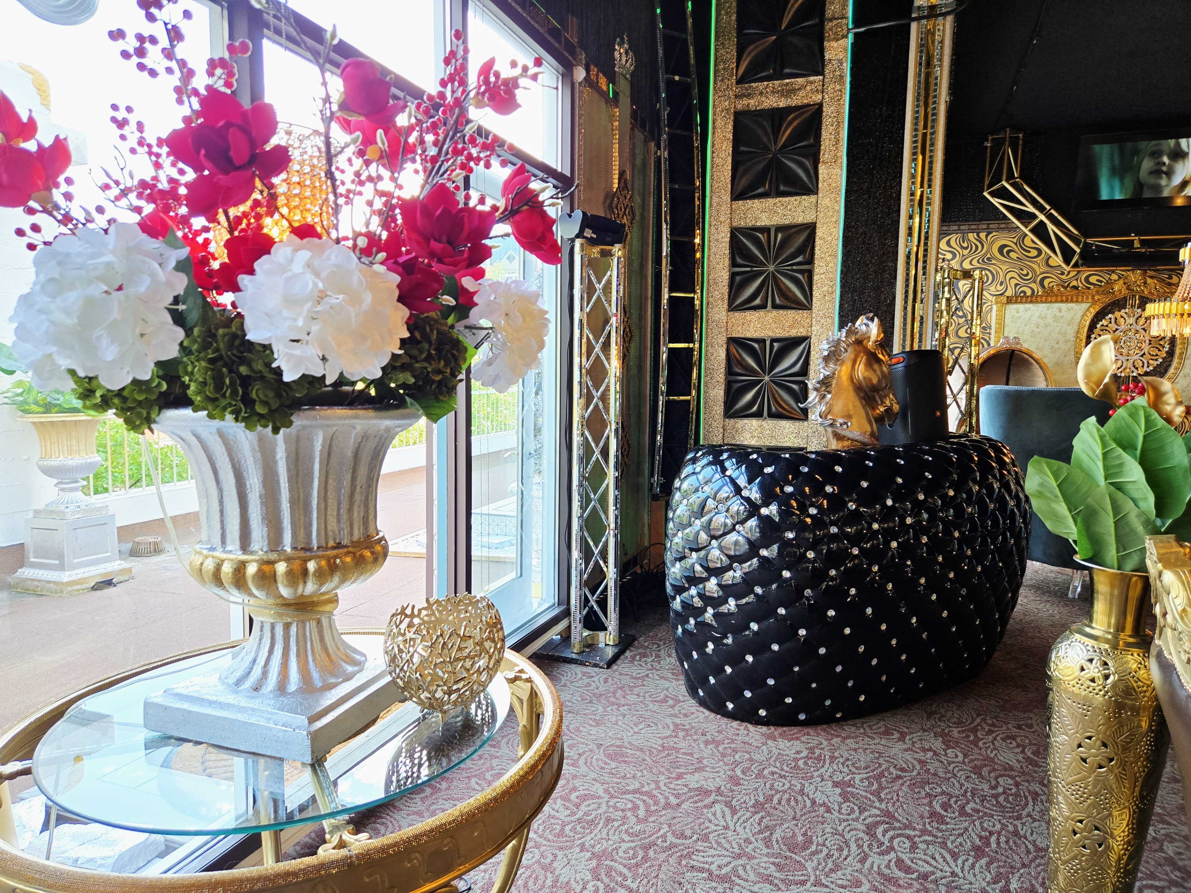 The image shows a stylish interior with decorative plants, a floral arrangement in a vase, and a large, black, studded furniture piece.