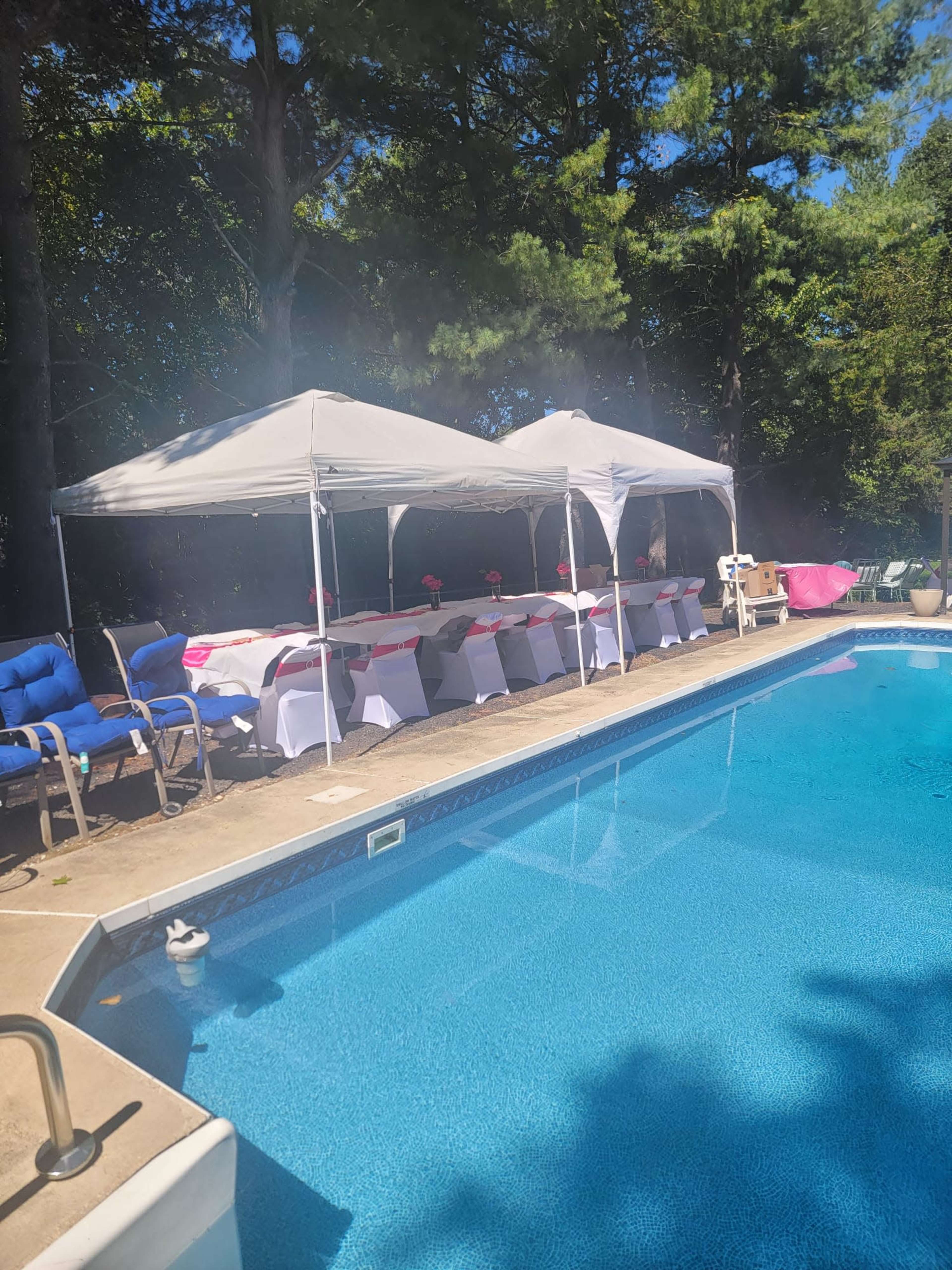 A swimming pool is surrounded by two white tents and tables set for a gathering, with blue lounge chairs nearby.