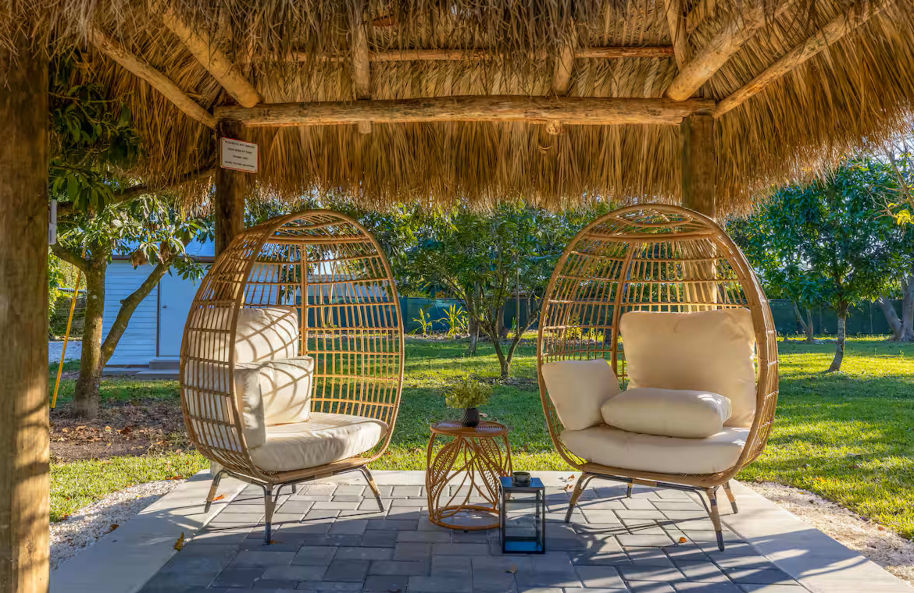 The image shows two woven chairs with cushions situated under a thatched roof, surrounded by greenery and a stone floor.