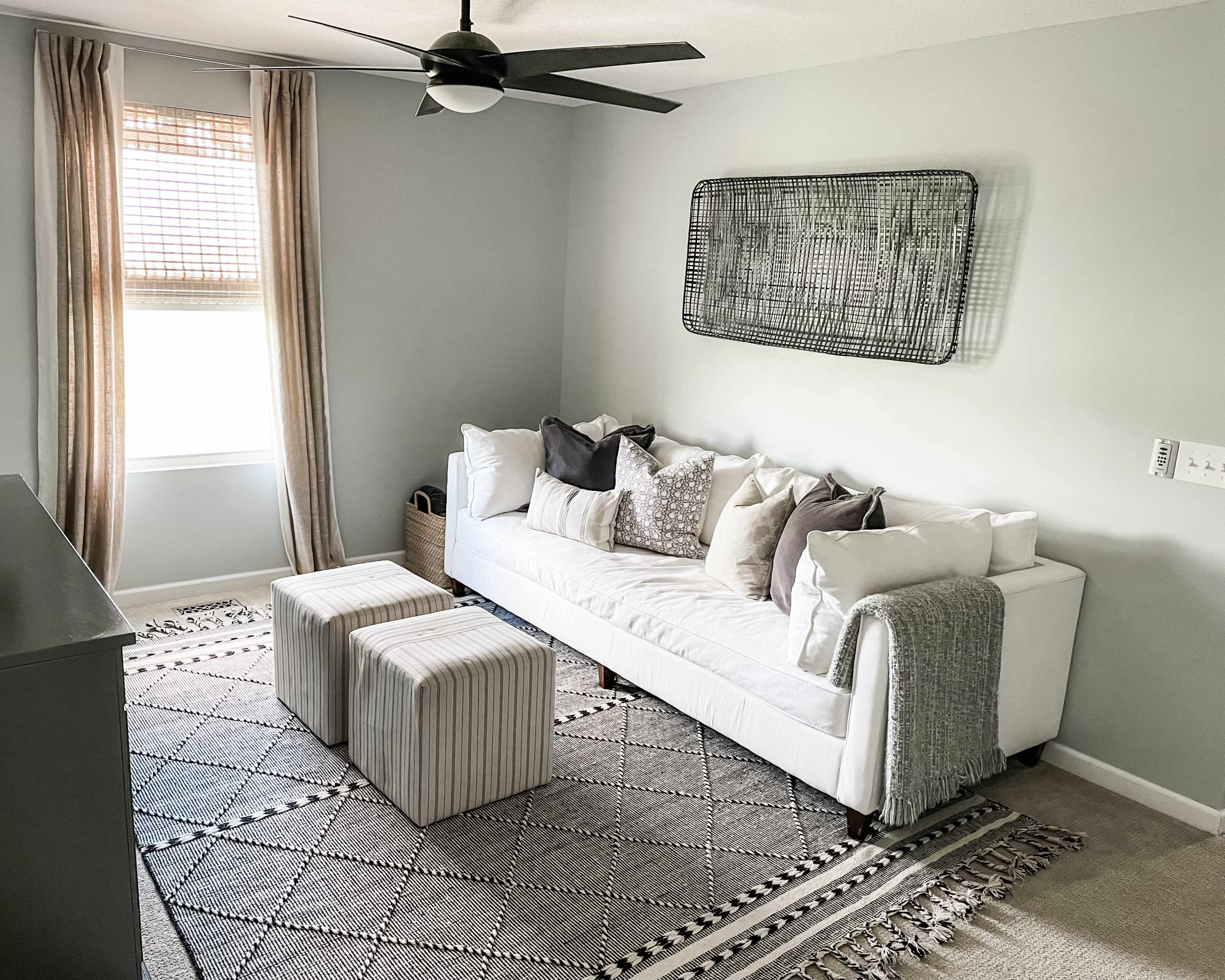 The room features a light-colored sofa adorned with various patterned cushions, two striped ottomans, and a geometric rug, all set against a neutral wall with a ceiling fan and window drapes.